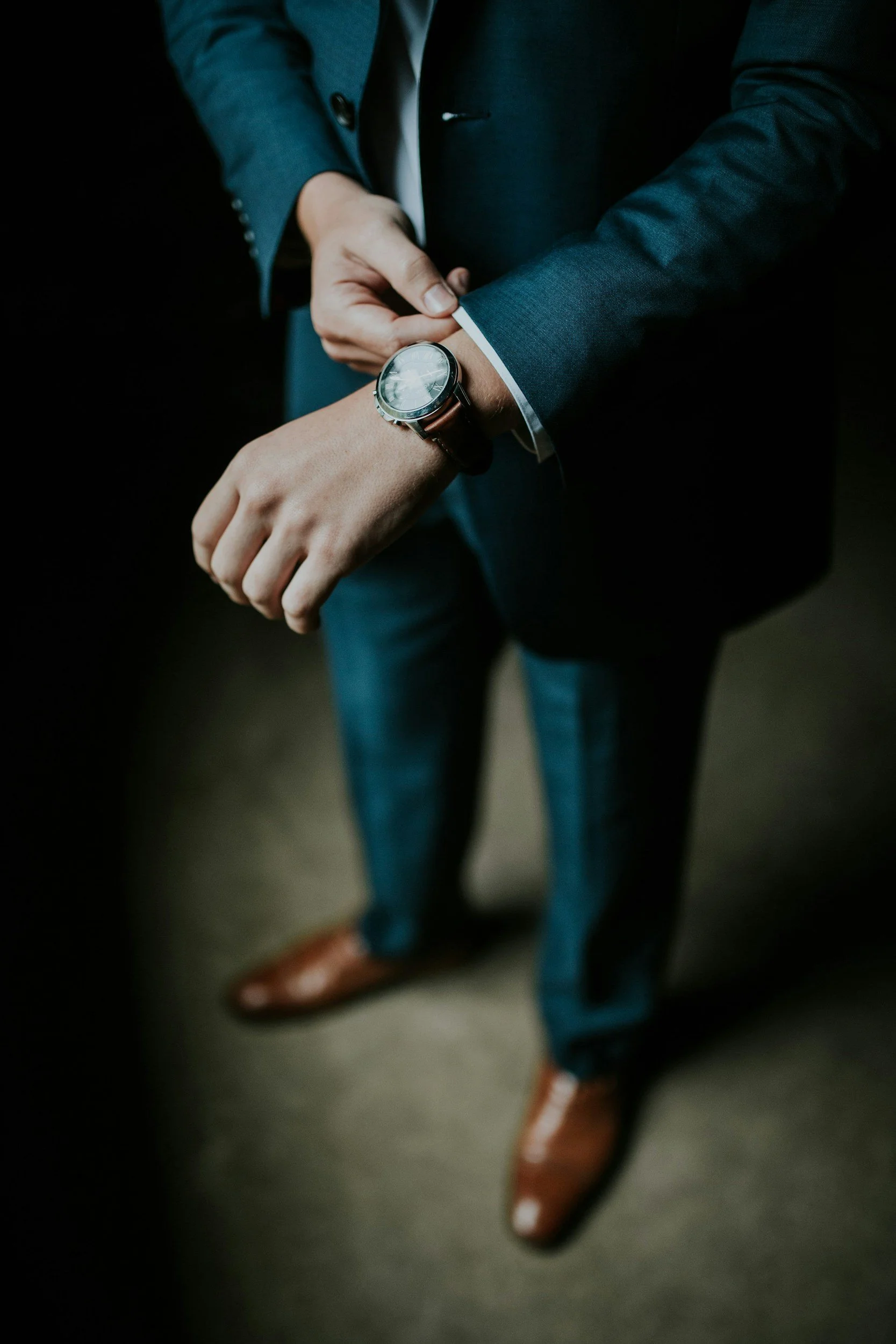 A man in a dark blue suit is adjusting his watch. He's wearing brown shoes and jeans.