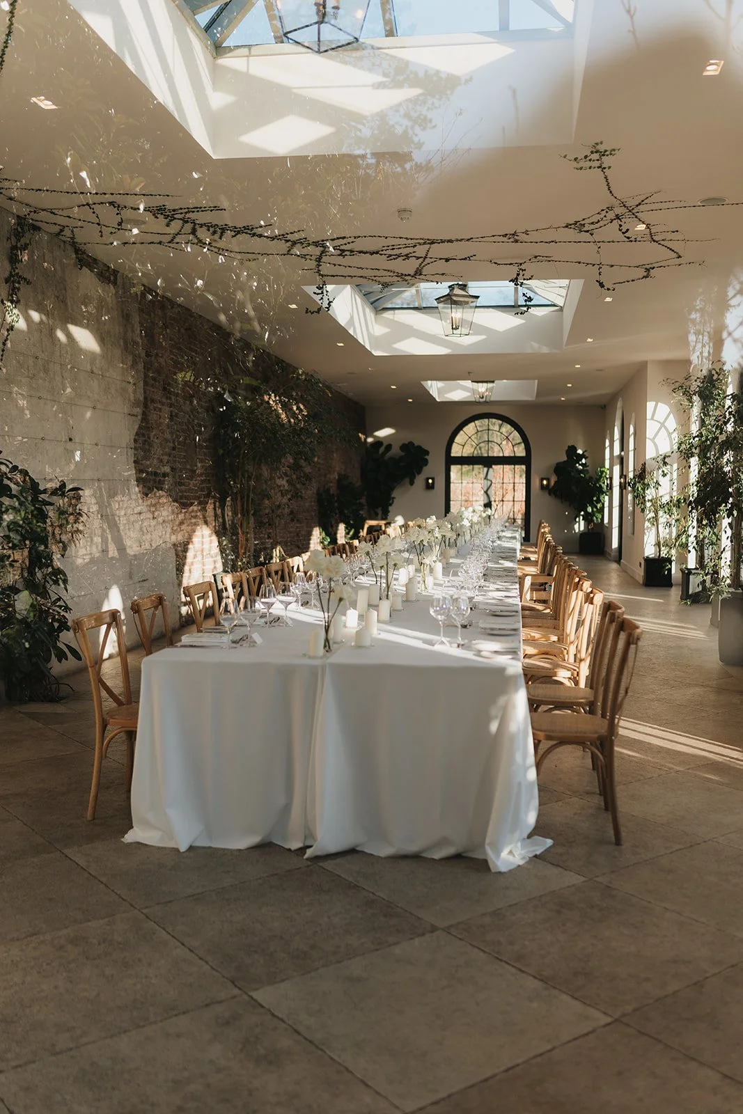 Long rectangular banquet table with white tablecloth, set with white flowers, candles, glassware, and place settings in a sunlit indoor event space with stone and brick walls, potted plants, and skylights.