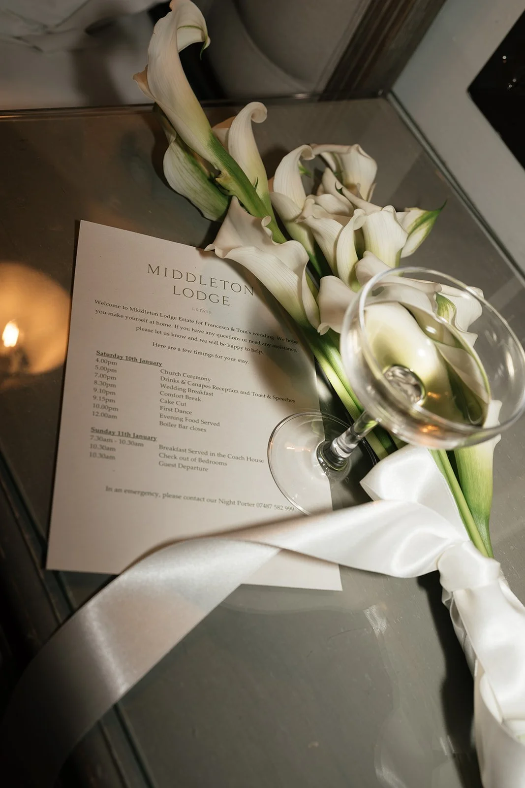 A bouquet of white calla lilies and a glass of wine placed on top of a printed wedding schedule at Middleton Lodge estate.