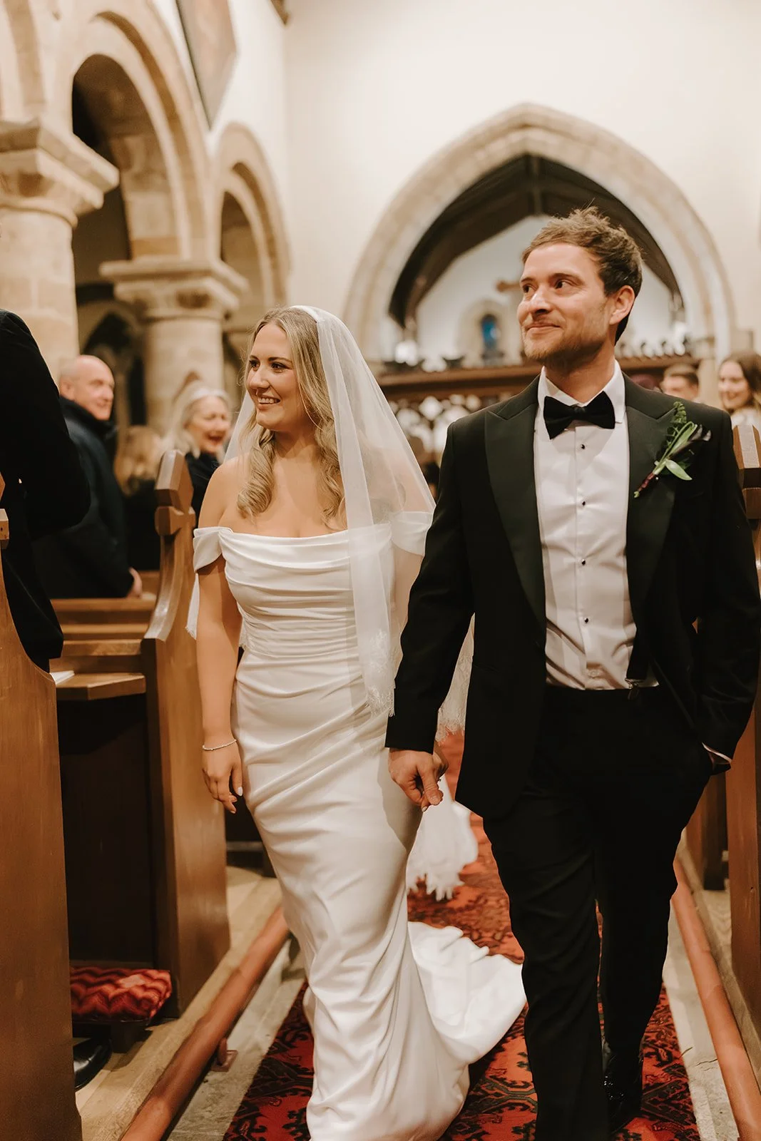 Bride and groom holding hands walking down the aisle inside a church with guests smiling in the background.