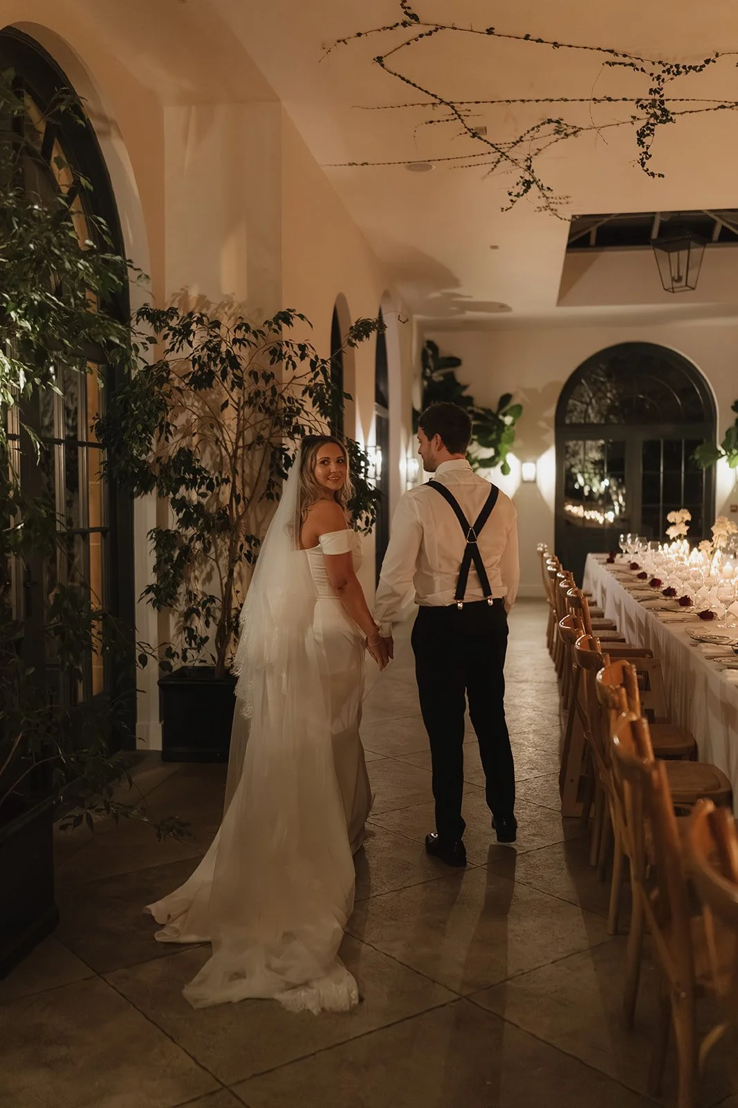 A bride and groom holding hands, standing on a lit dance floor in a wedding reception hall. The bride is wearing a white wedding gown with off-the-shoulder sleeves and a veil, looking back at the camera and smiling. The groom is dressed in white shir