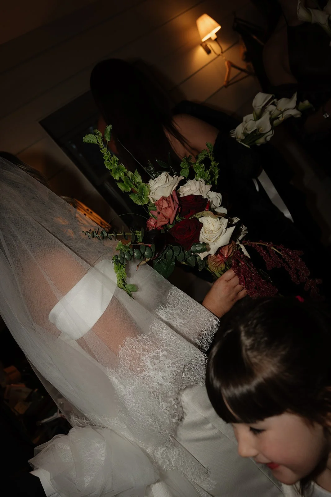 A bride wearing a lace gown and veil holding a bouquet of roses, with a woman in black and a young girl nearby, in a warmly lit room.