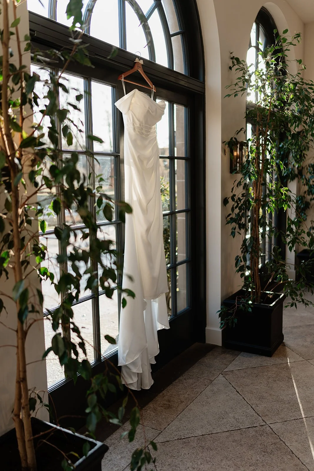 A white wedding dress hanging on a wooden hanger on a window frame in a well-lit room with large glass windows, surrounded by green indoor plants.