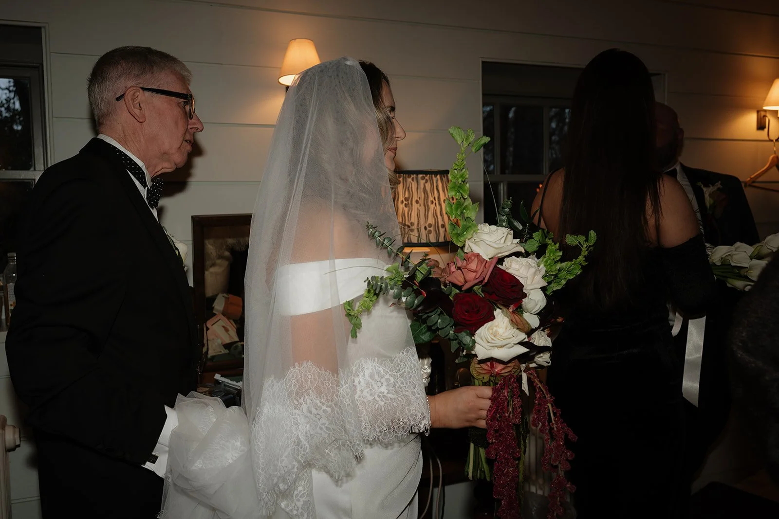 Bride holding a large bouquet of flowers at a wedding ceremony, standing with her wedding gown and veil, indoors with warm lighting.