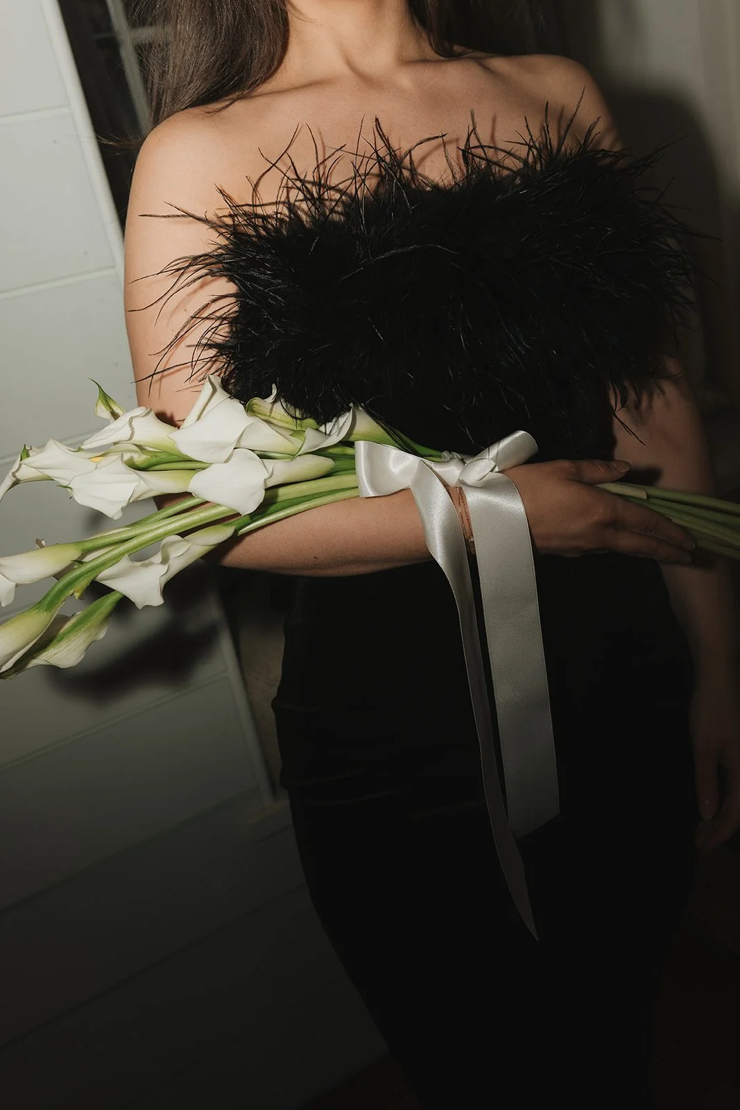 A woman wearing a black feathered top, holding a bouquet of white calla lilies wrapped with a white ribbon.