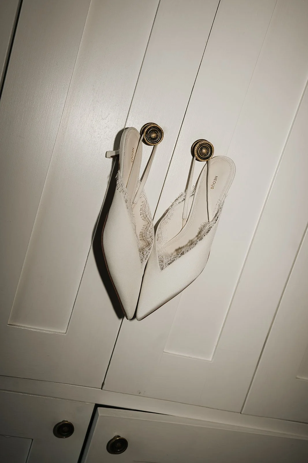 Pair of white high-heeled shoes with pointed toes hanging on a white cabinet door by decorative gold knobs.
