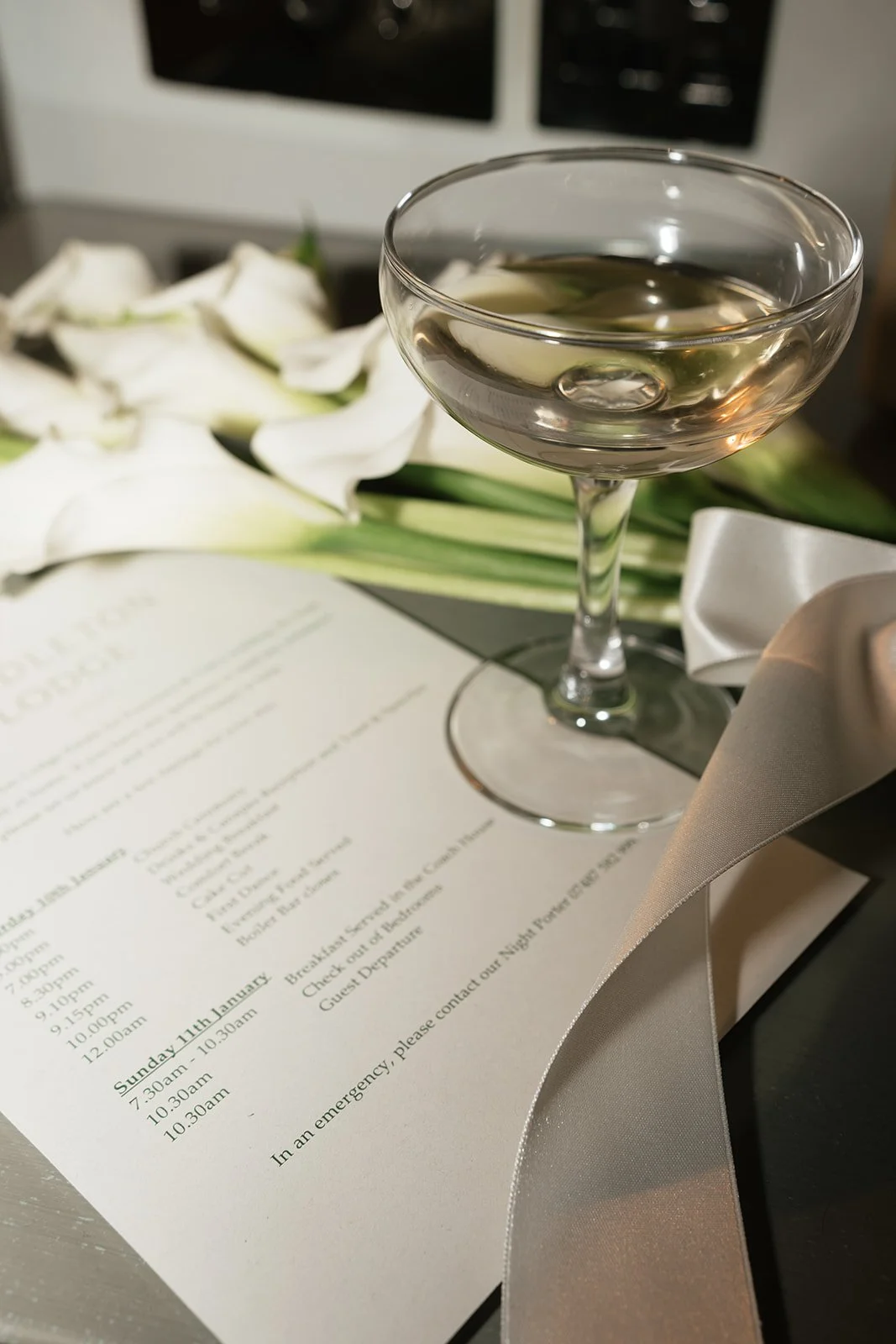 A cocktail glass filled with a clear beverage, placed on a table next to a menu, white calla lilies, and a satin ribbon.