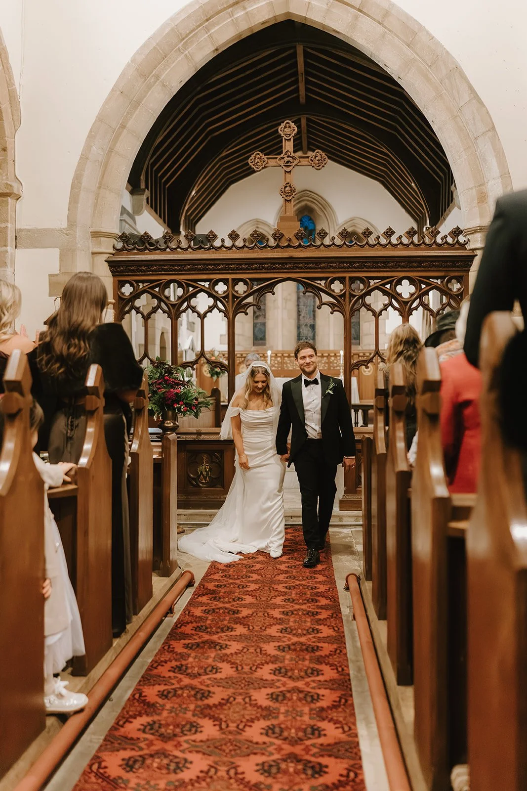 A bride and groom walking down the aisle inside a church, the bride wearing a white wedding dress and veil, the groom in a black tuxedo. Guests are seated on wooden pews, watching and smiling. The church has high arched ceilings, stone walls, and woo
