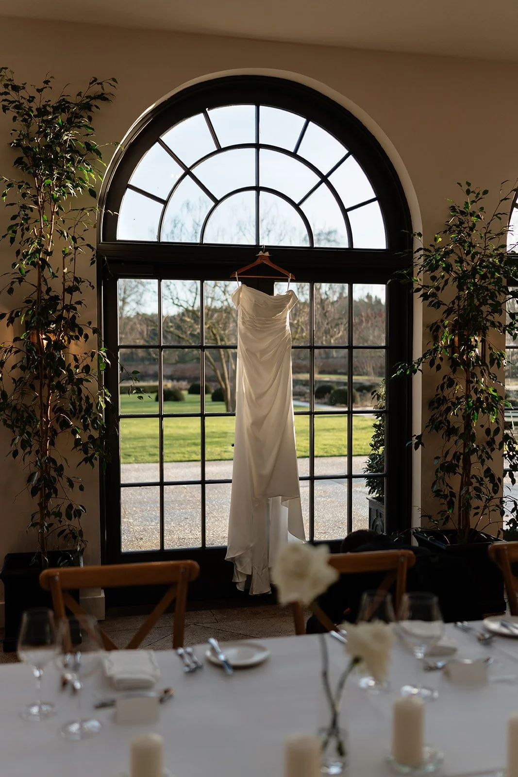 A white wedding dress hanging on a hanger in front of a large arched window with trees and a grassy yard outside.
