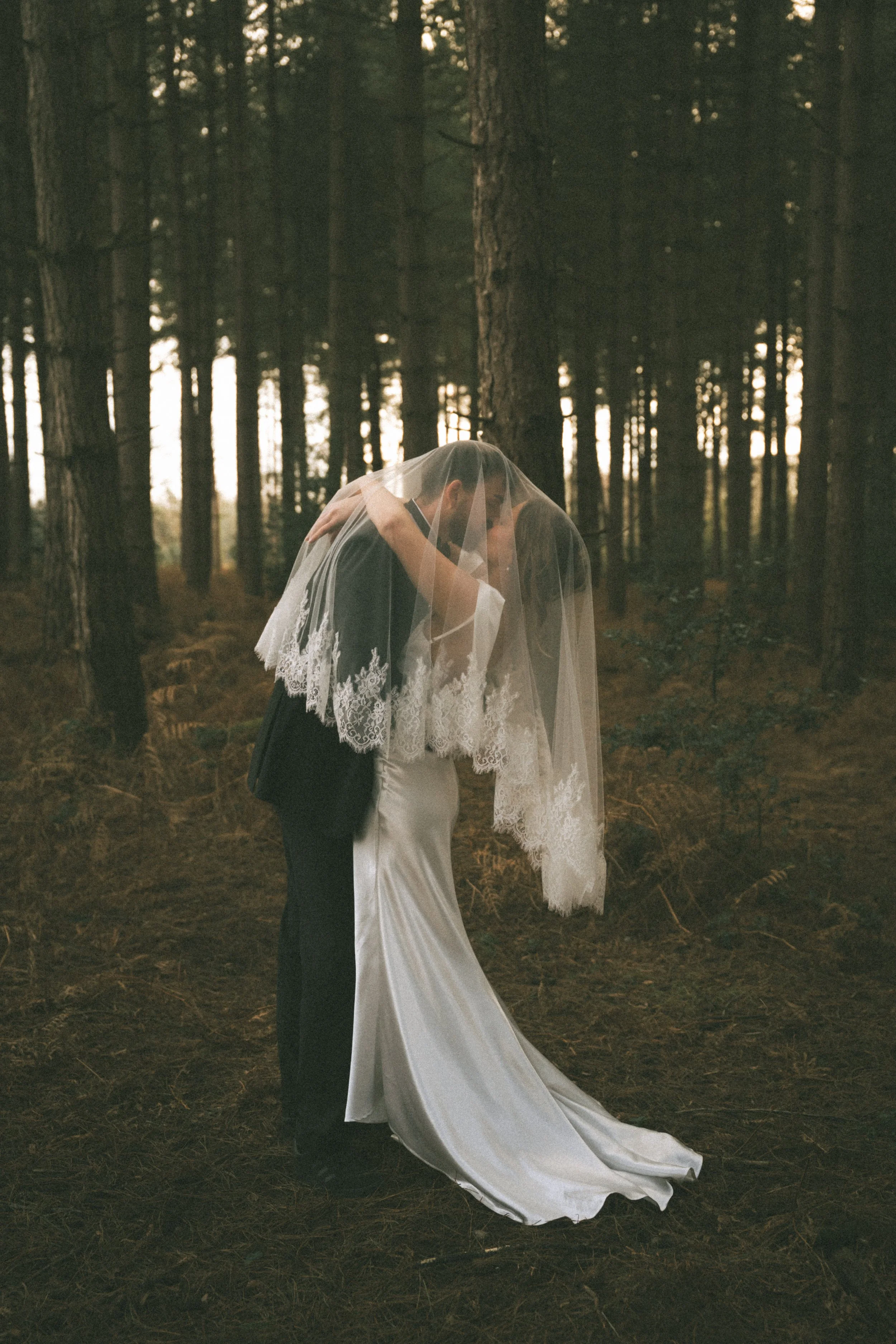 A bride and groom sharing a kiss in a forest, with the bride wearing a lace wedding veil and a satin wedding dress.