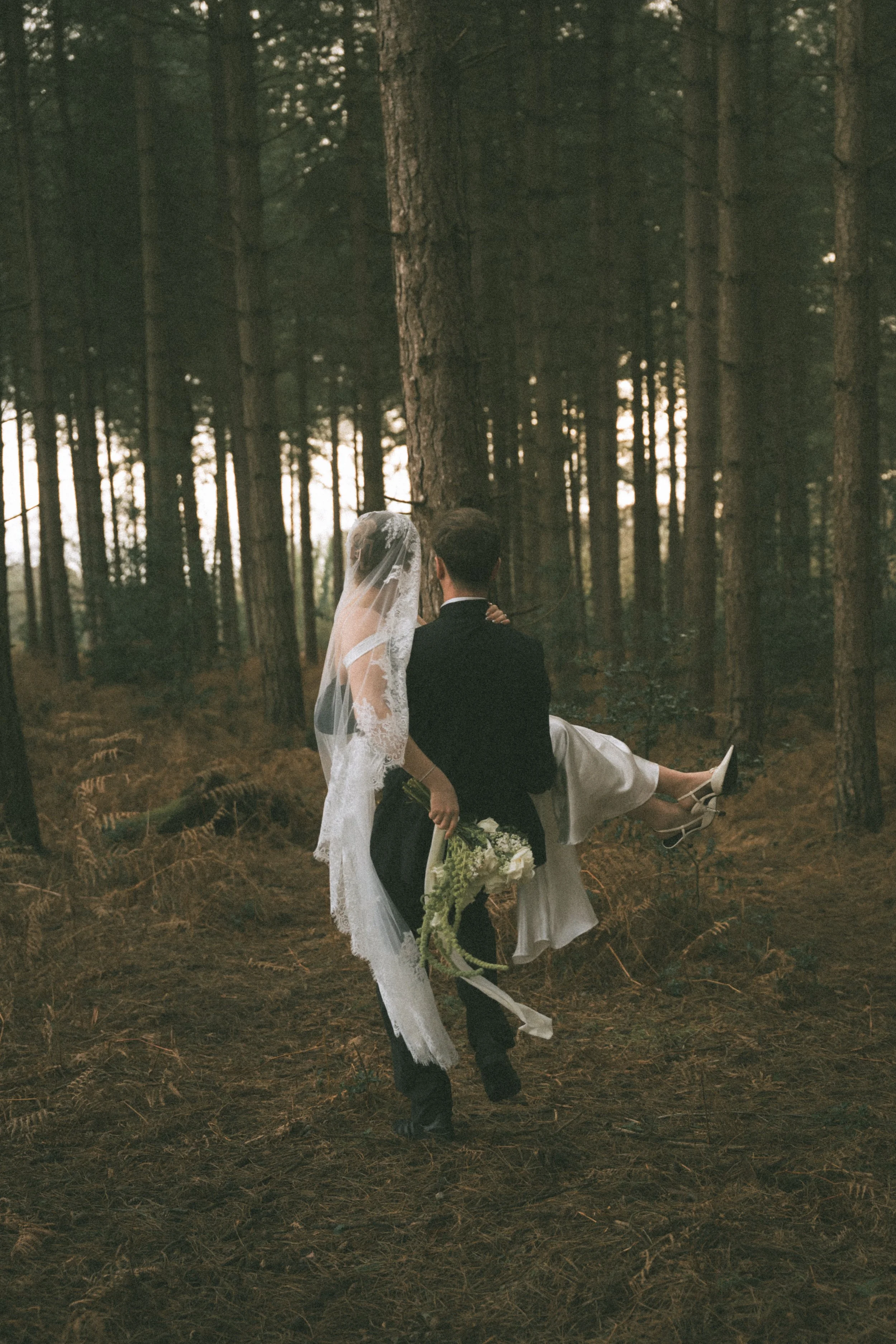 A bride in a wedding dress with a veil being carried by a groom in a suit through a forest with tall trees for a Norfolk Wedding by Editorial Luxury Wedding Photographer by Lauren Elise Photography