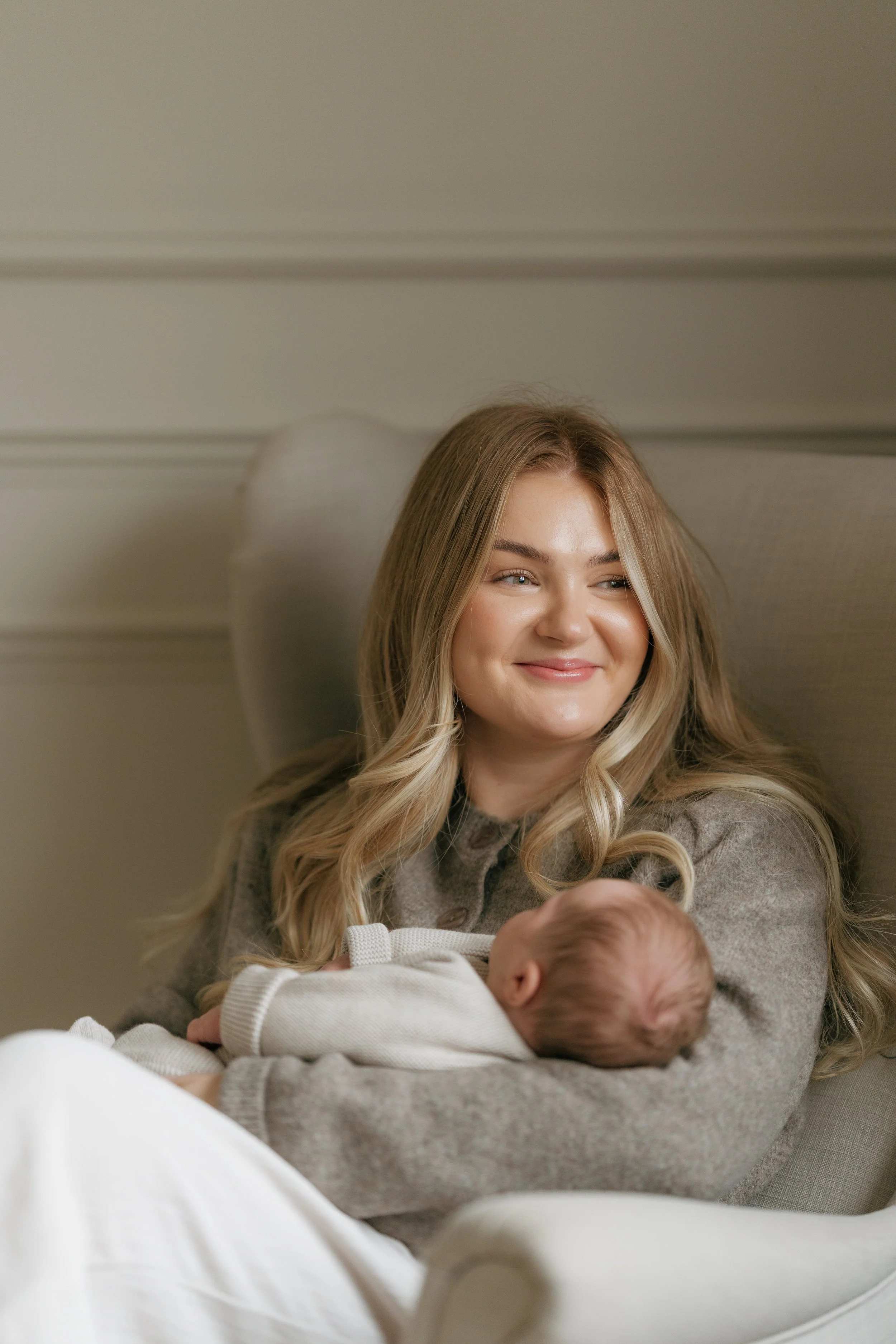 A young woman with long blonde hair sitting on an armchair holding a sleeping baby, both smiling softly.