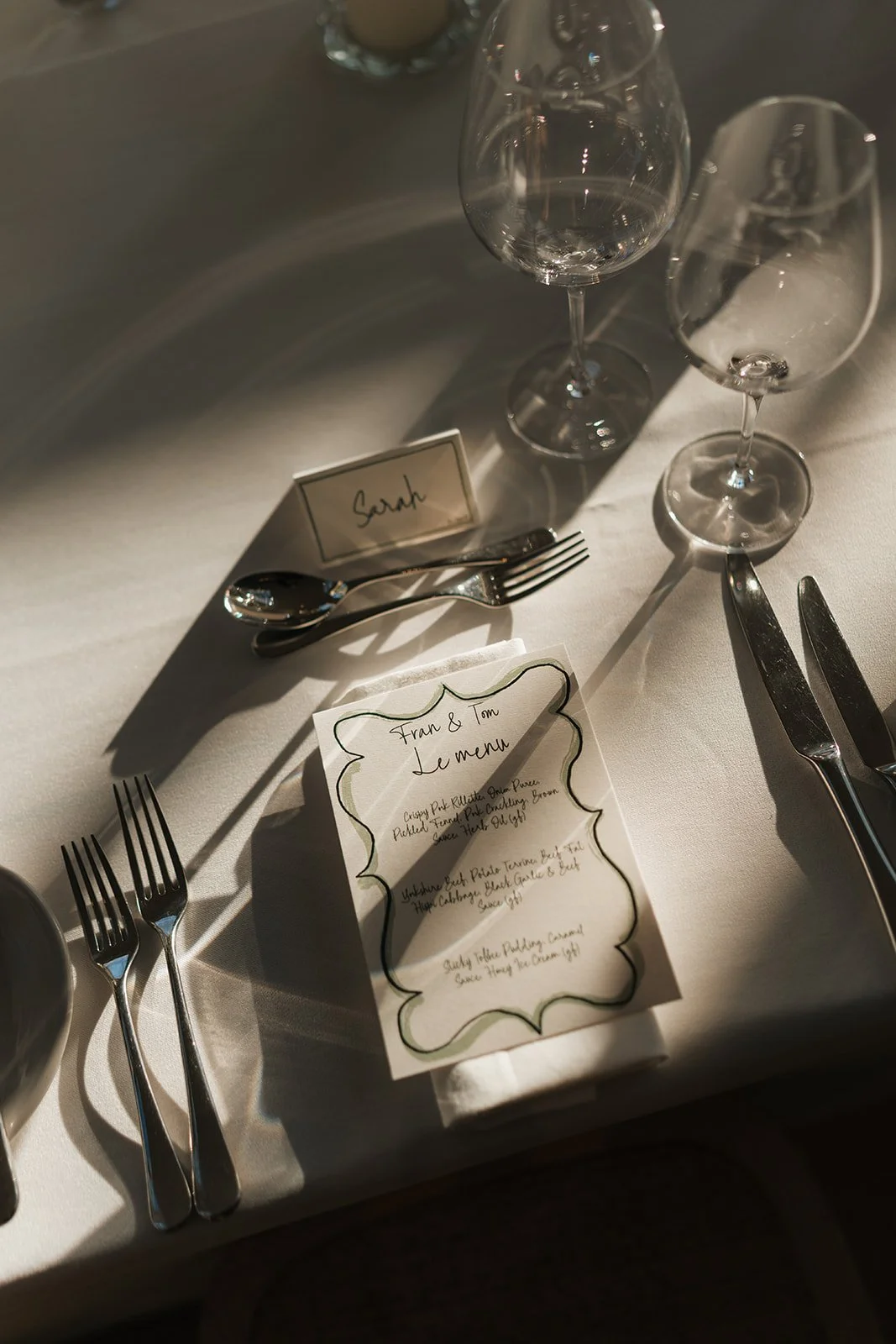 A formal dining table set with two wine glasses, plates, forks, knives, spoons, a folded napkin, a table card with the name 'Sarah', and a printed menu card for Fran & Tom.