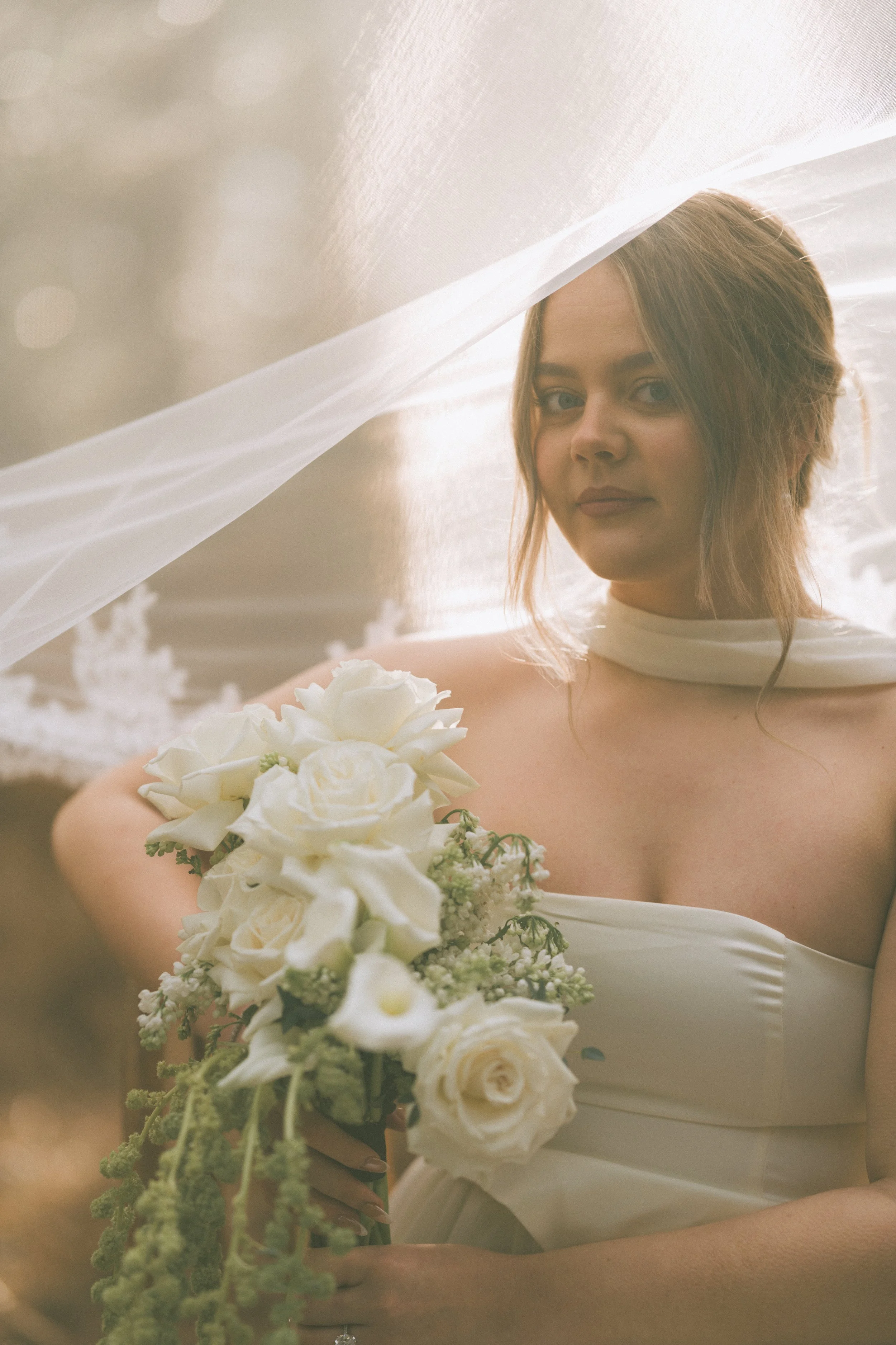 A woman in a white dress holding a bouquet of white flowers, with a sheer veil draped over her head, outdoors during daylight for a Norfolk Editorial Luxury Wedding
