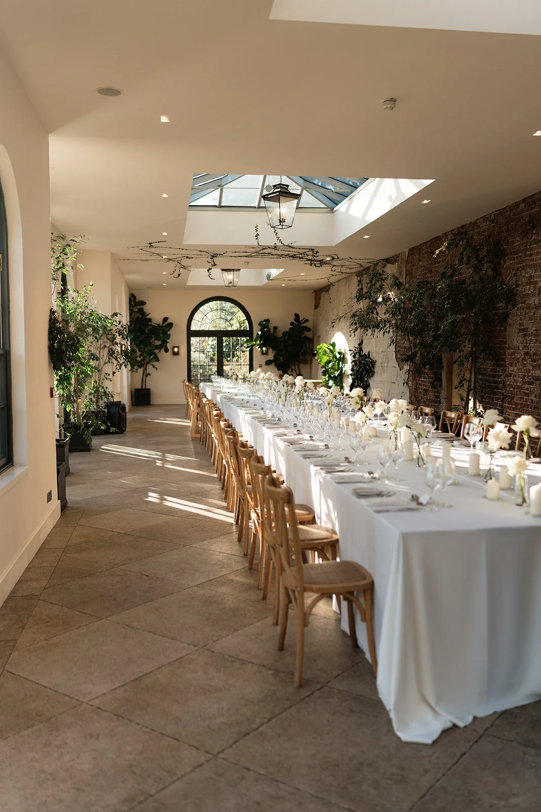 Long dining table set with white tablecloth, glassware, and floral centerpieces in a bright, decorated event space with plants and skylights.