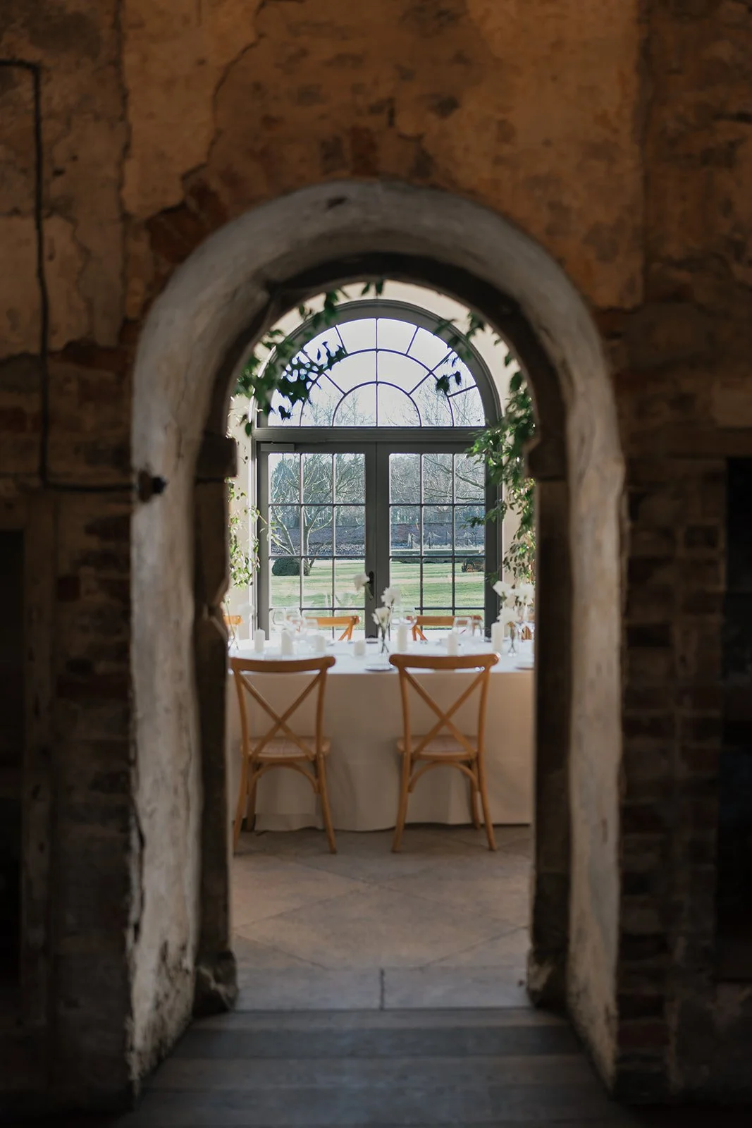 A view through a stone archway into a bright room with a window, a table set for a meal, and wooden chairs, overlooking a lawn and trees outside.