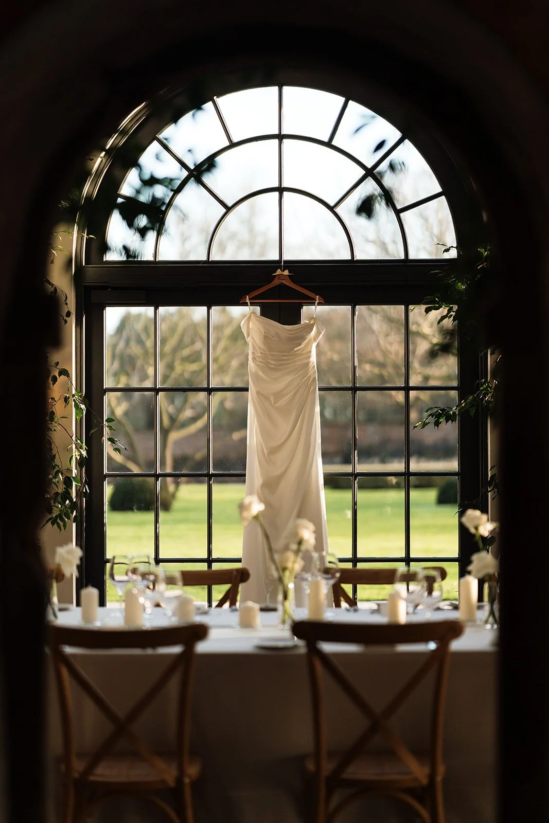 A white wedding dress hanging on a wooden hanger in front of a large window with a semi-circular top, with a decorated table with candles and floral arrangements below.