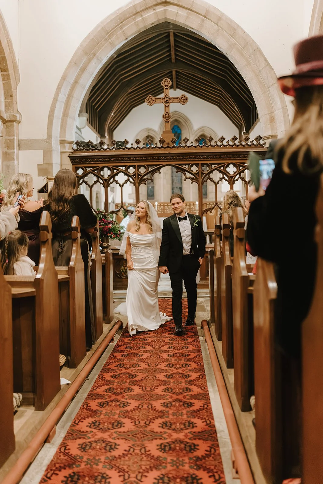 A bride and groom walking down the aisle in a church, surrounded by guests taking photos and celebrating their wedding.