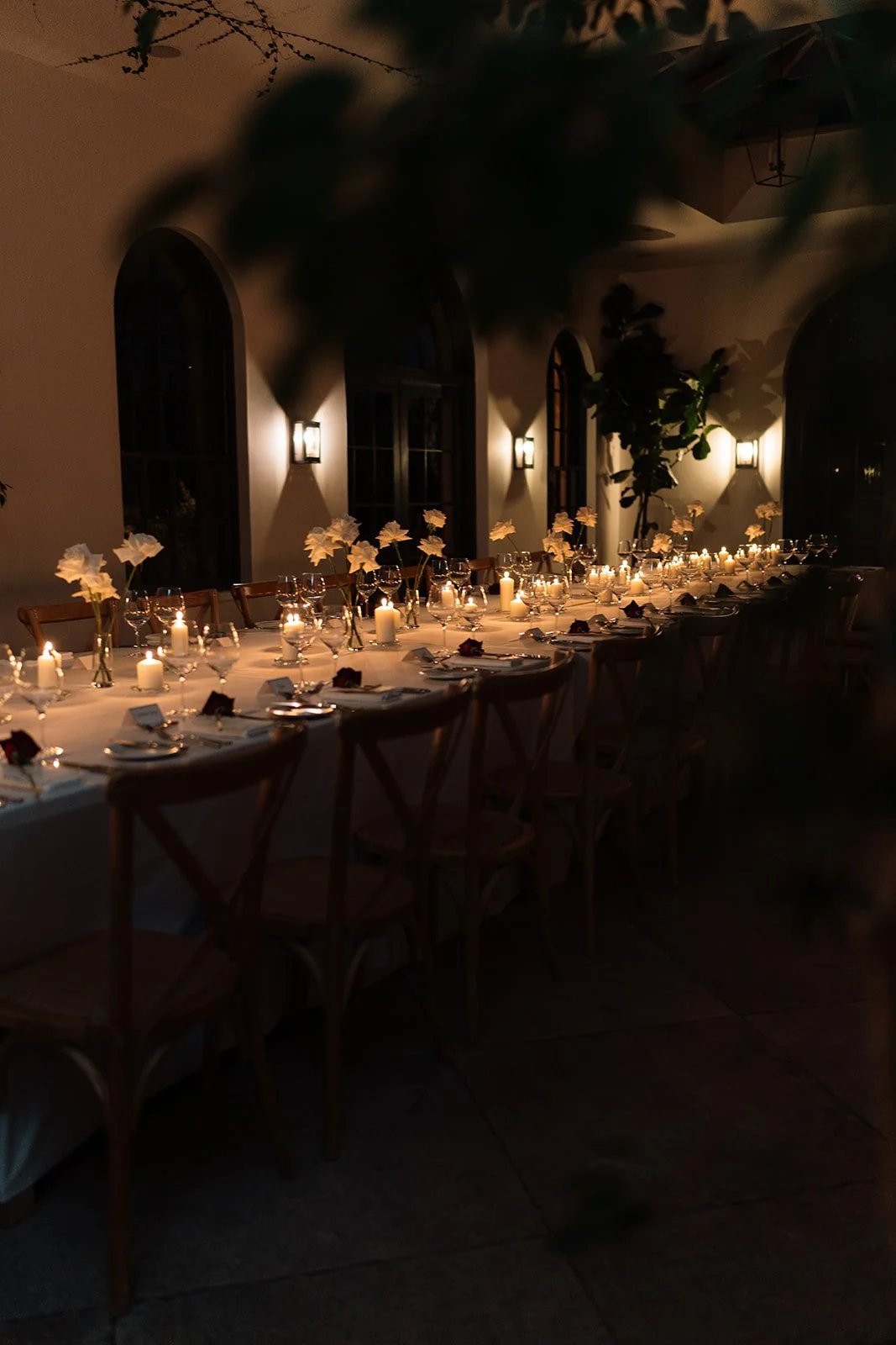 A long dining table set for a formal event, decorated with candles, flowers, and place settings, in a dimly lit room with arched windows and wall sconces.