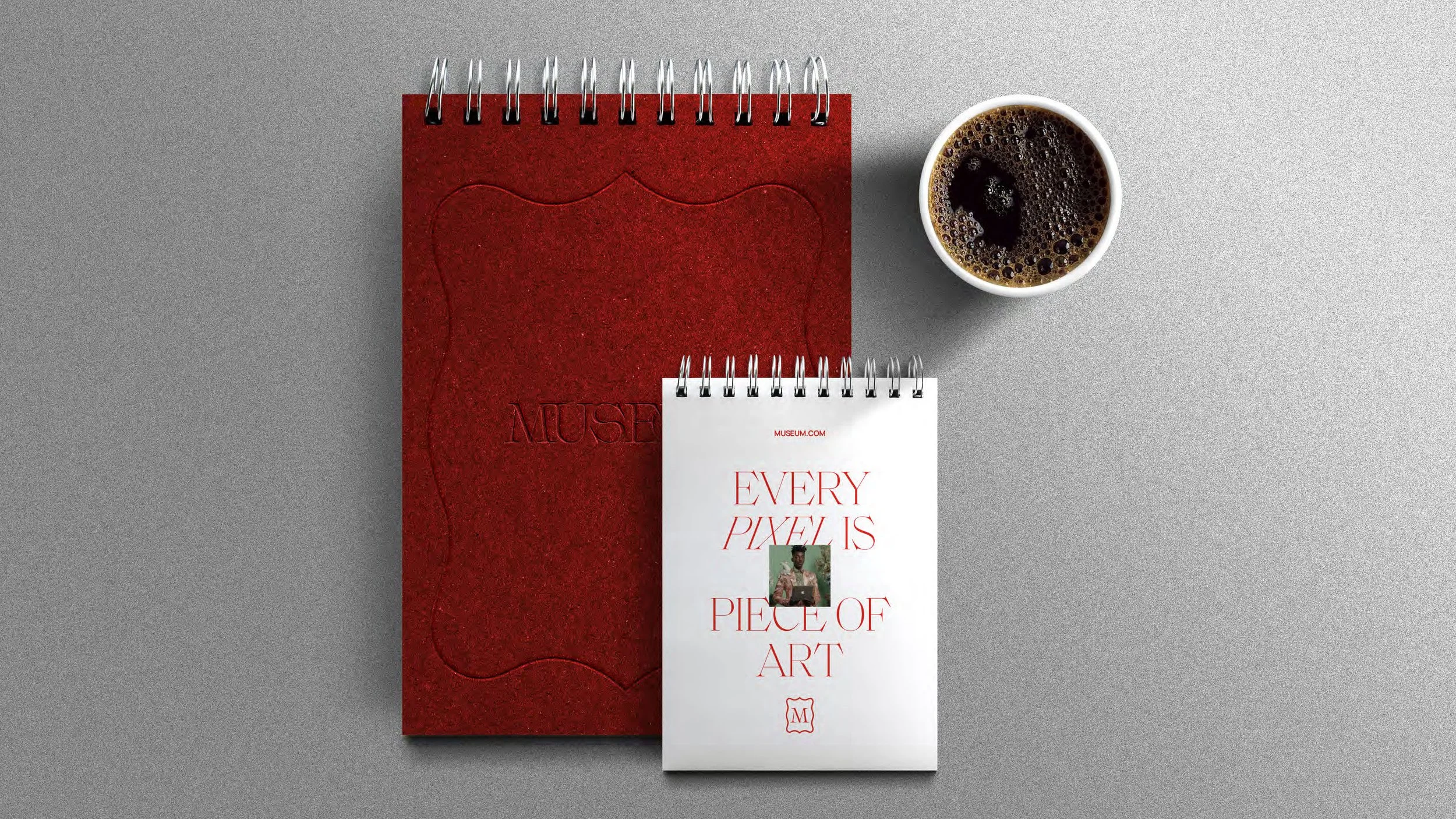 Flat lay of a red sketchbook with embossed design, a white spiral-bound notepad with red text reading 'Every Pixel Is Piece of Art' and a small image, and a cup of coffee on a gray surface.