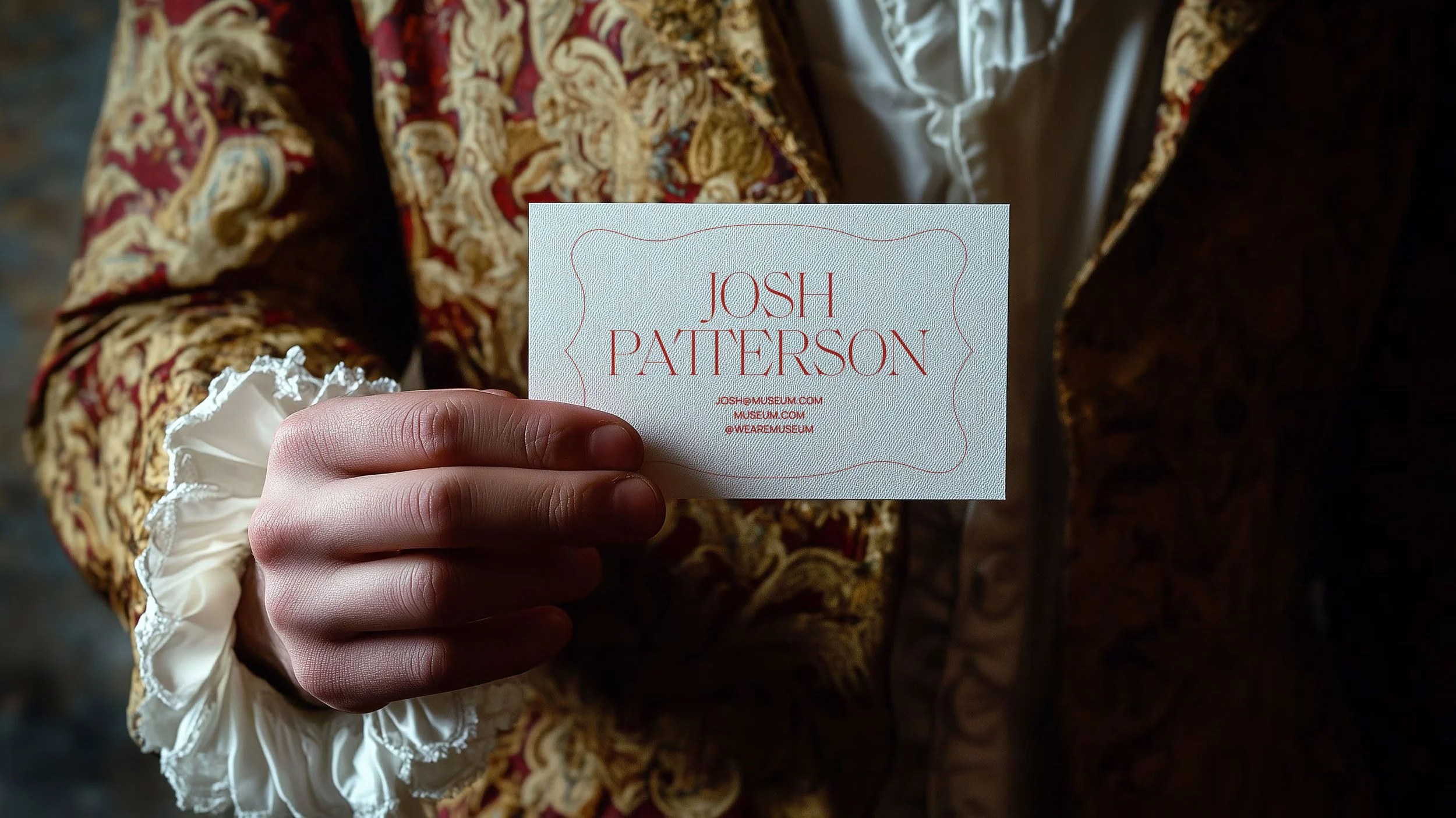 A person dressed in an ornate, patterned jacket holds a business card for Josh Patterson, with the person's hand and part of their clothing visible.