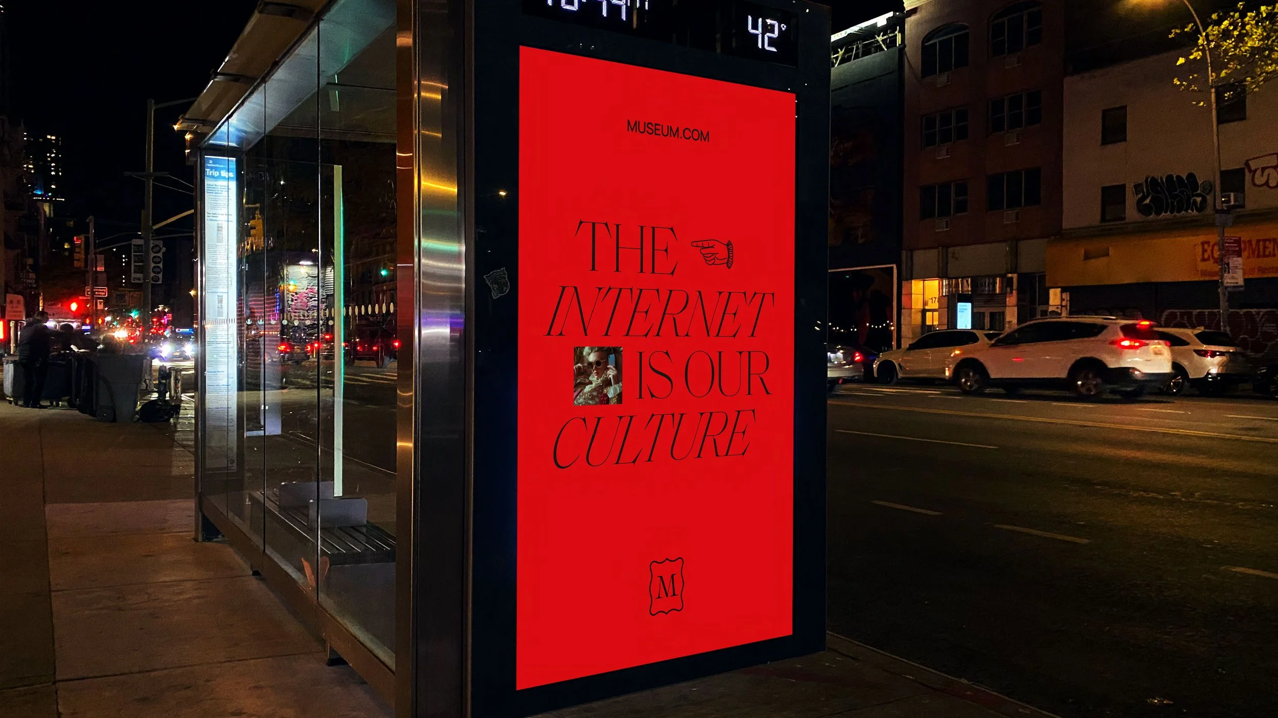 A nighttime city street scene with a digital billboard at a bus stop displaying a red advertisement for a museum, stating 'The Internet is Our Culture' with a small image of an astronaut and some decorative elements.
