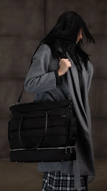 A woman with black hair wearing a gray coat, holding a large black bag, standing against a dark background.