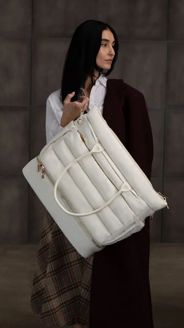 Woman holding a large white handbag, dressed in a white shirt and a plaid skirt, with a dark coat draped over her shoulder, standing against a dark wall background.