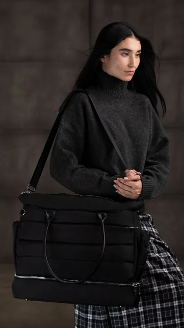 A woman with long dark hair wearing a black turtleneck sweater carrying a large black bag and wearing plaid pants.