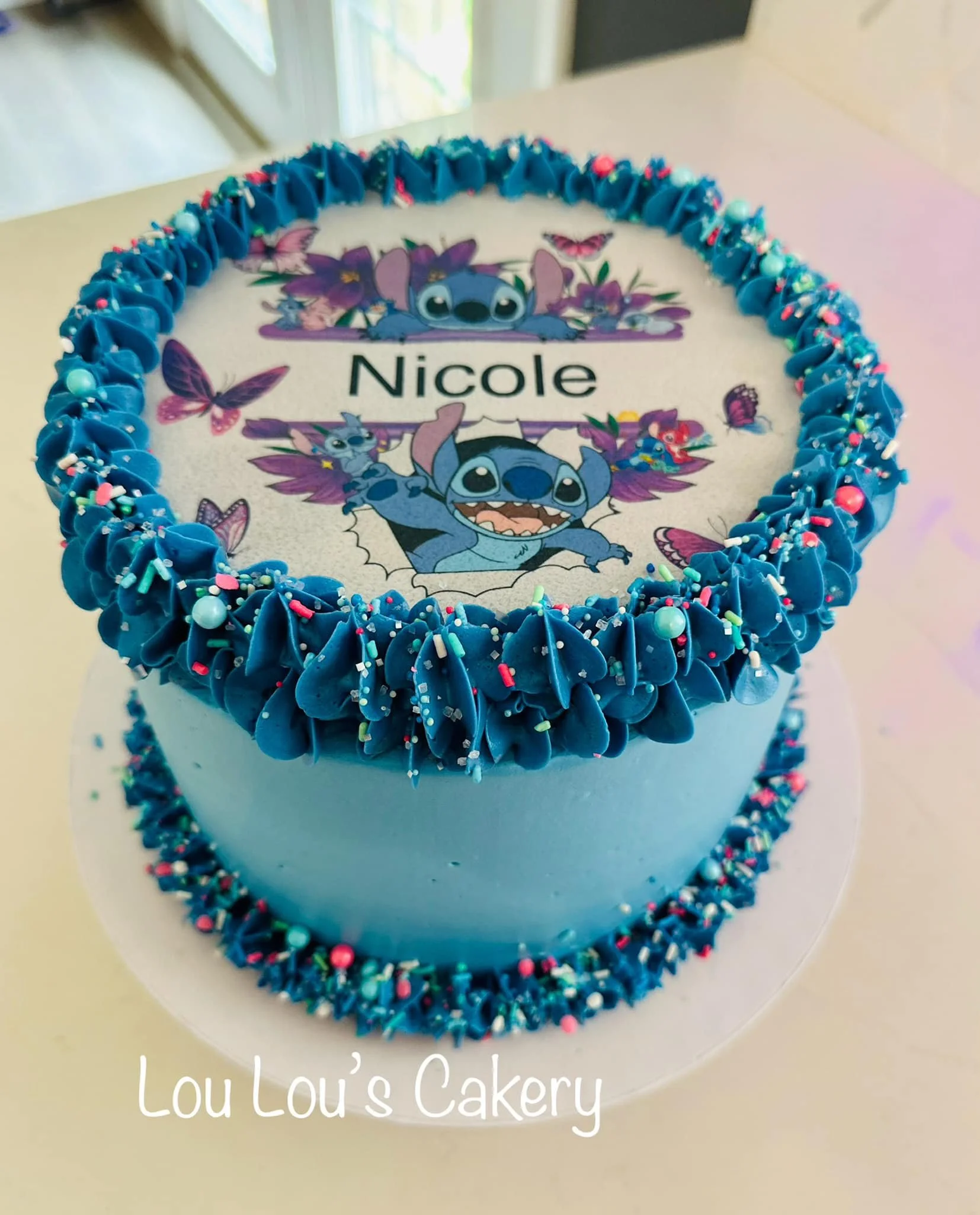 Blue birthday cake with an 'Lilo & Stitch' themed edible image on top, featuring Stitch and butterflies, with pink, blue, and white sprinkles around the edges, and the name 'Nicole' in the center. The cake is from Lou Lou's Cakery.