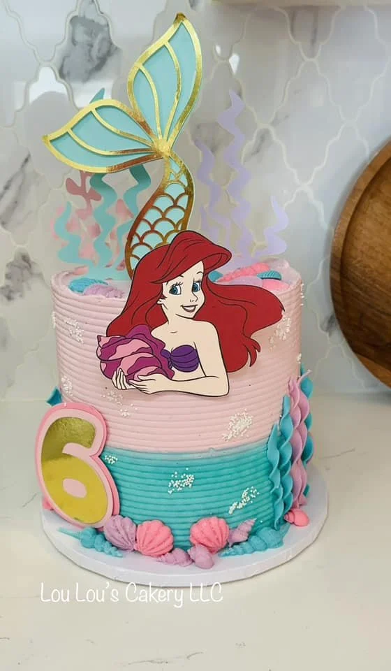 A birthday cake decorated with a 'The Little Mermaid' theme, featuring a large image of Ariel with red hair and purple seashell top, a mermaid tail topper, and a large yellow number 6 candle.