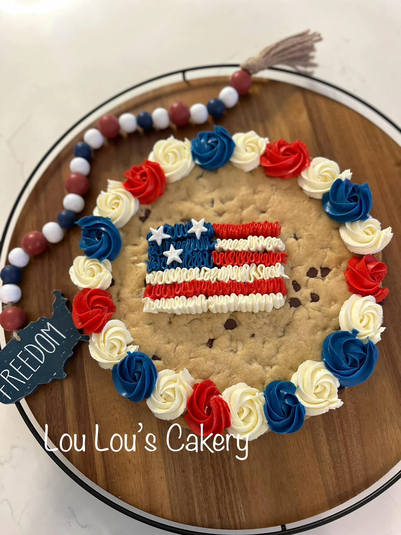 Decorated large cookie with a frosting American flag in the center, surrounded by red, white, and blue frosting roses, on a wooden board. There is a small flag that says "FREEDOM" to the side, and the text "Lou Lou's Cakery" at the bottom.
