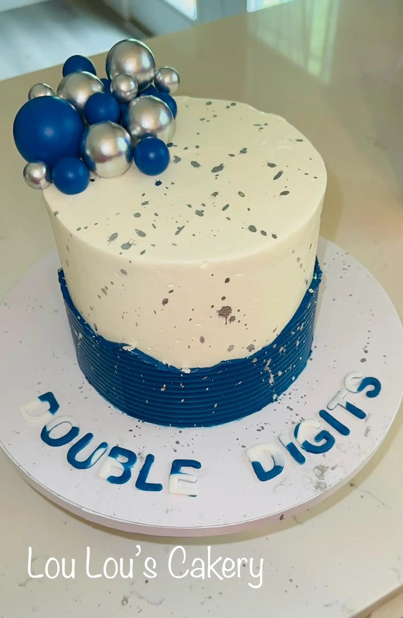 A two-tiered cake with a dark blue bottom tier and a speckled white top tier, decorated with silver and dark blue spherical cakes on top, with the words "DOUBLE DIGITS" on the cake board.