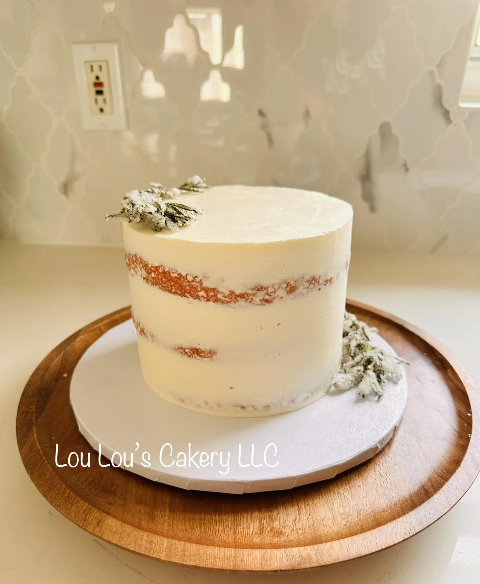 A round layered cake with white buttercream frosting, decorated with frosted lavender sprigs on top, displayed on a wooden board. The cake has red streaks inside, visible through the frosting.