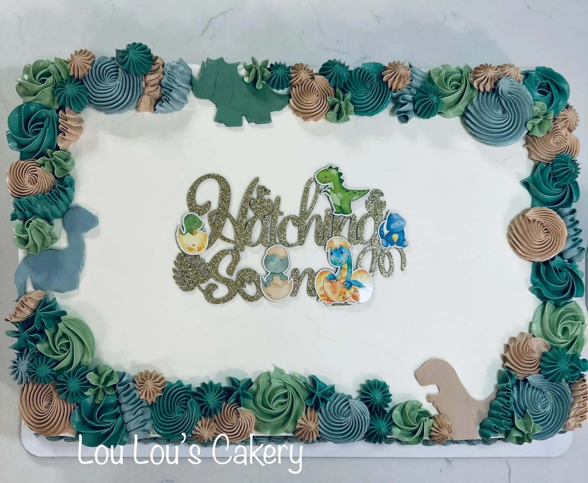 A rectangular celebration cake decorated with pastel-colored buttercream swirls and rosettes in shades of blue, green, and beige, featuring dinosaur-themed decorations and a glittery gold cake topper that reads 'Happy Datchiqua Son' with dinosaur sti