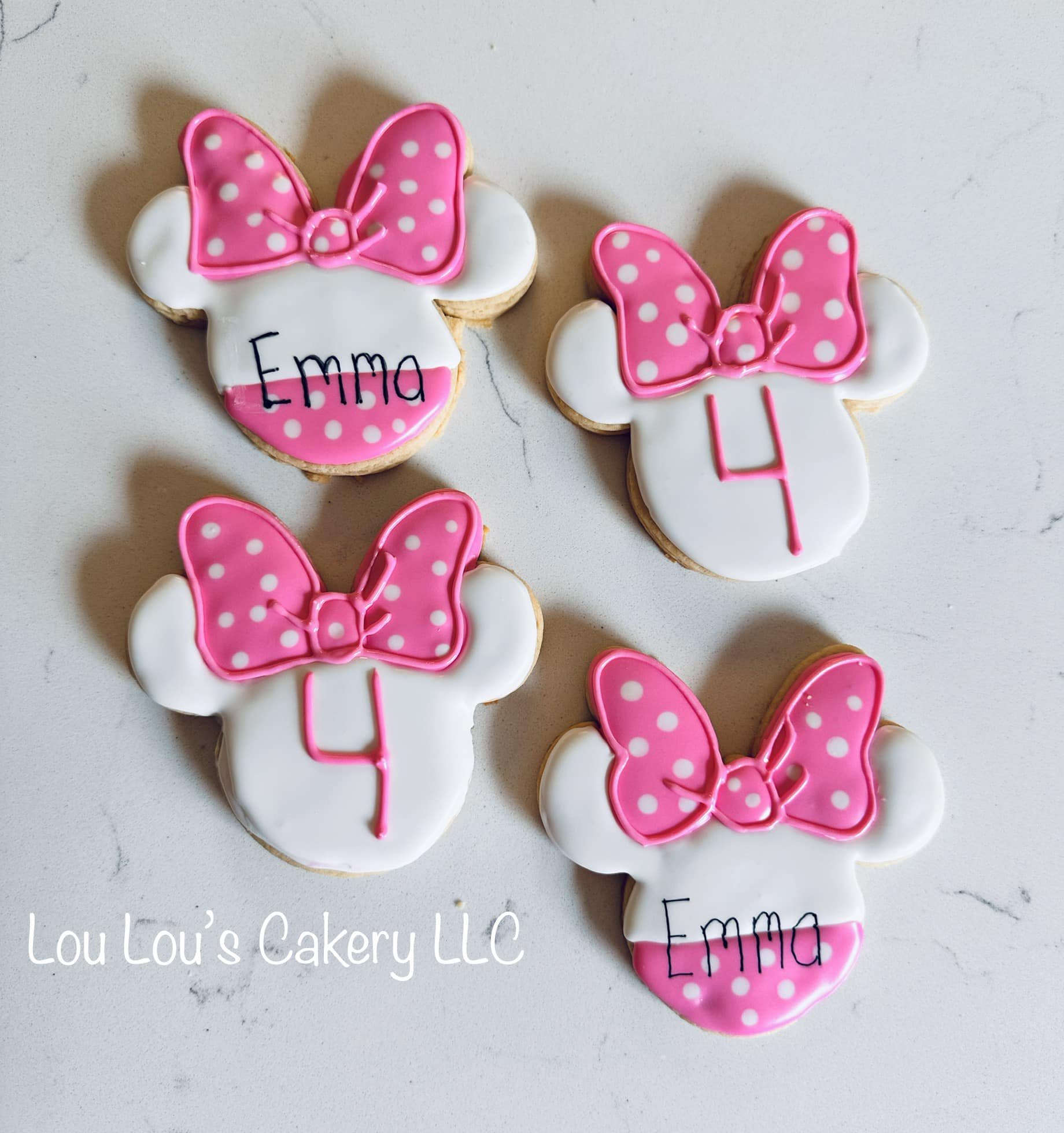Decorative cookies in the shape of a Minnie Mouse head with a pink polka dot bow, white face, and a pink number 4 on the chin, some cookies have the name Emma written on them, on a white surface with Lou Lou's Cakery LLC written in the bottom left co