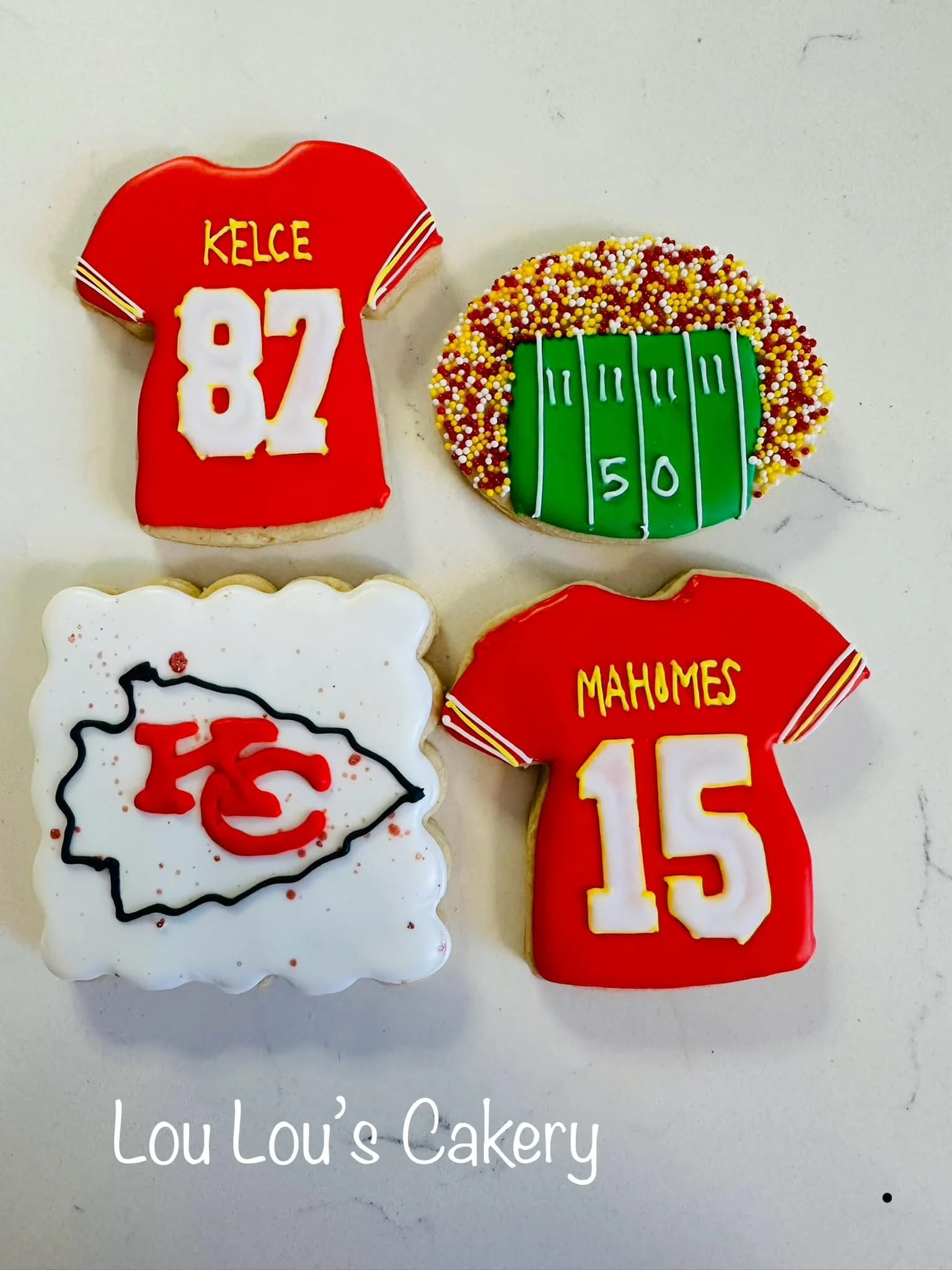 Decorative cookies with sports themes and Kansas City Chiefs logos, shaped like jerseys and a football field, with text 'Kelce 82' and 'Mahomes 15' and the Chiefs logo, on a white surface, with a caption 'Lou Lou's Cakery'.