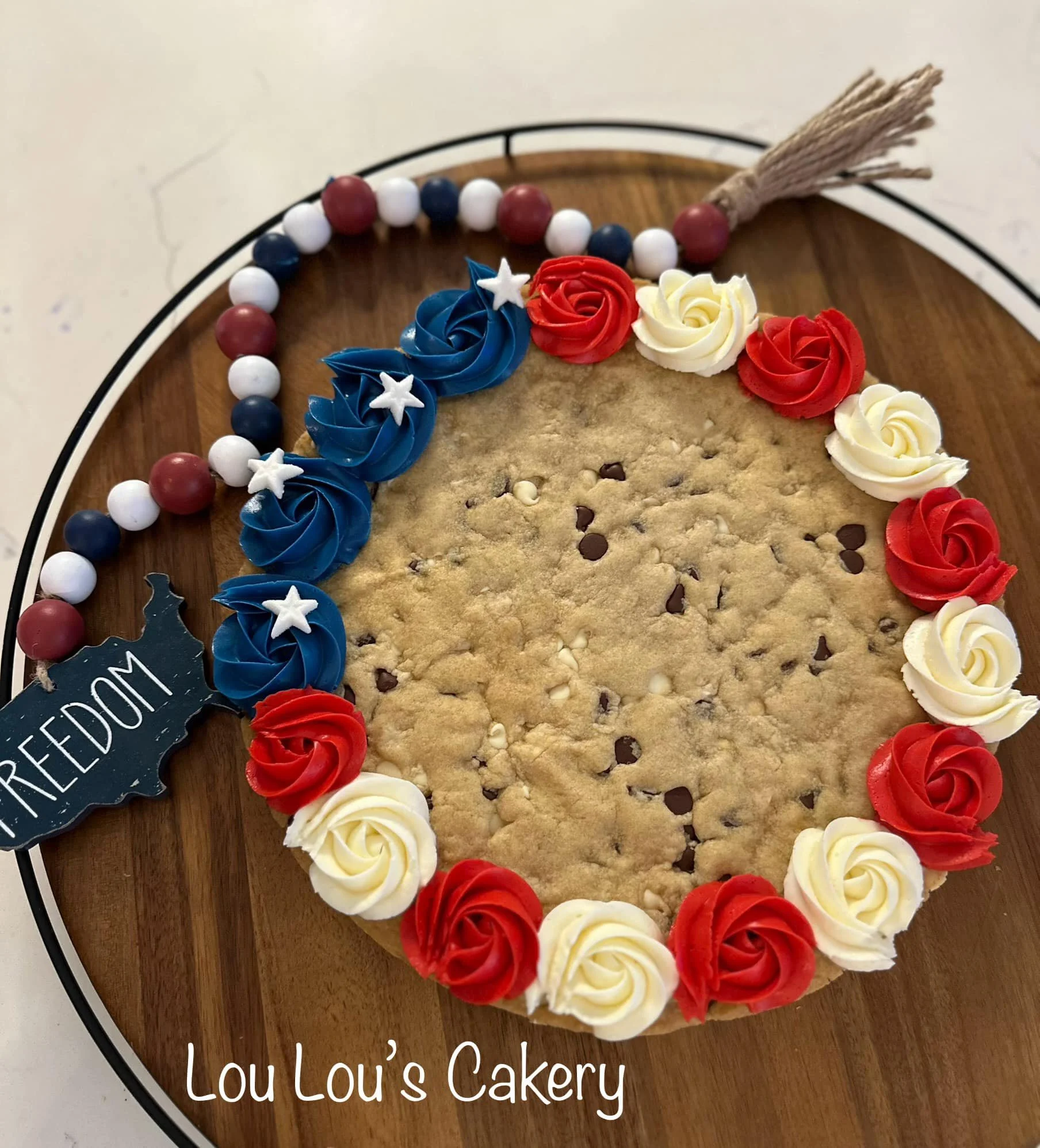 Decorated cookie cake with red, white, and blue icing roses and stars, a beaded border, and a tag that says 'FREEDOM', on a wooden board with GPIO logo, labeled 'Lou Lou's Cakery'.