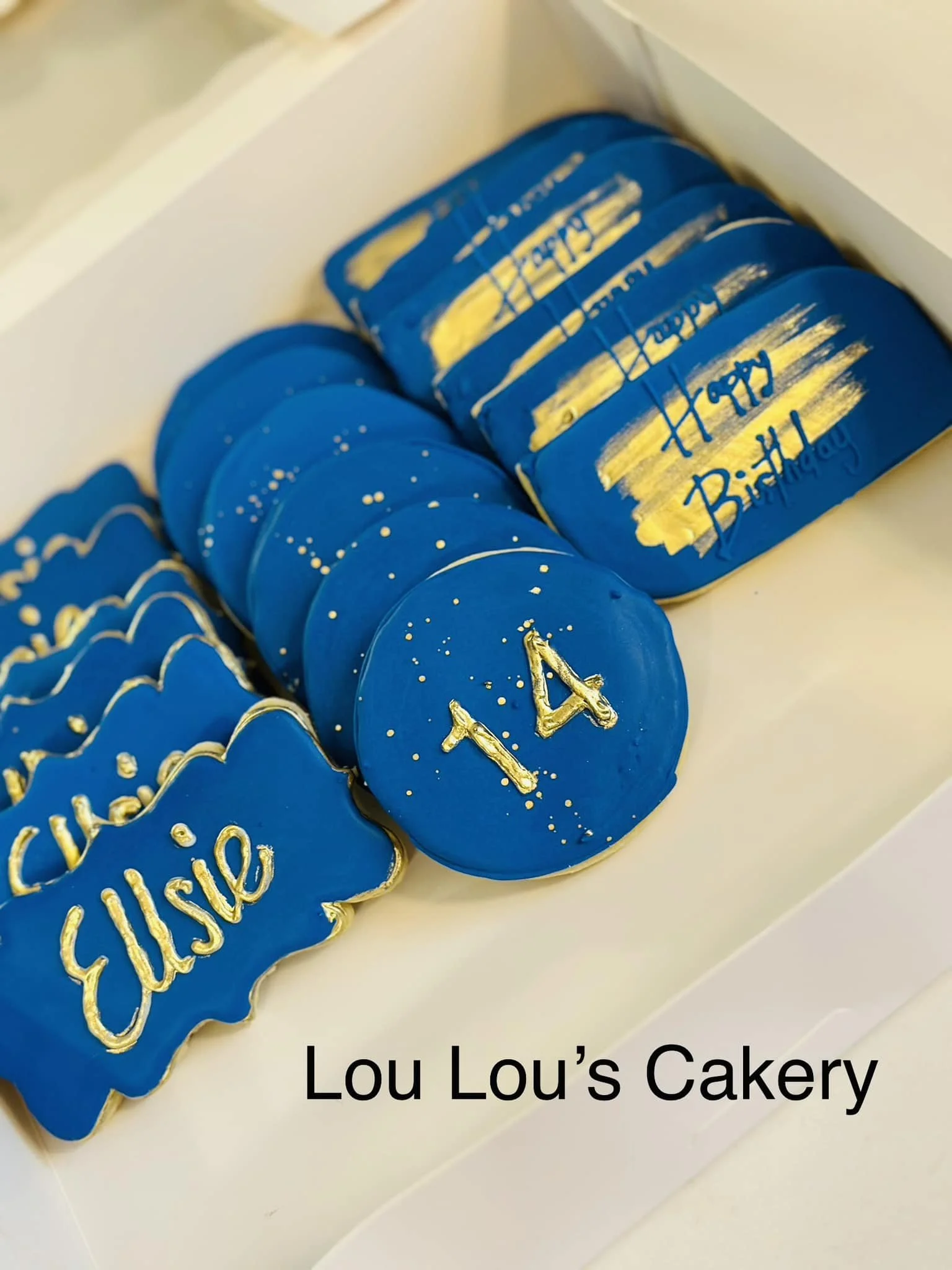 Blue birthday cookies with gold writing and designs in a box, including a cookie with the number 14 and another with the name Elsie.