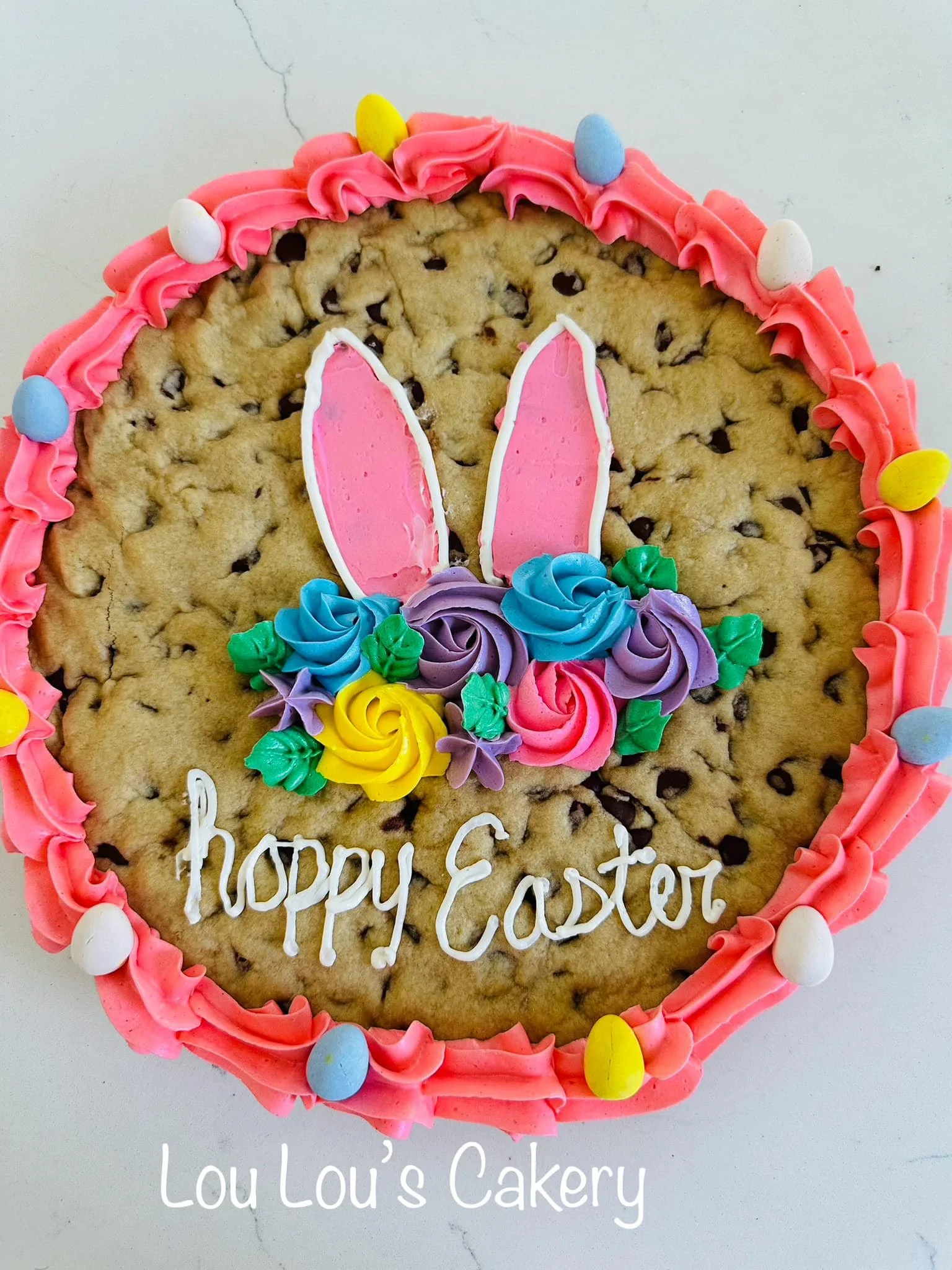 Easter cookie decorated with pink icing, colorful sprinkles, and a bunny face with pink and white ears, with a message that says "happy Easter" and "Lou Lou's Cakery".