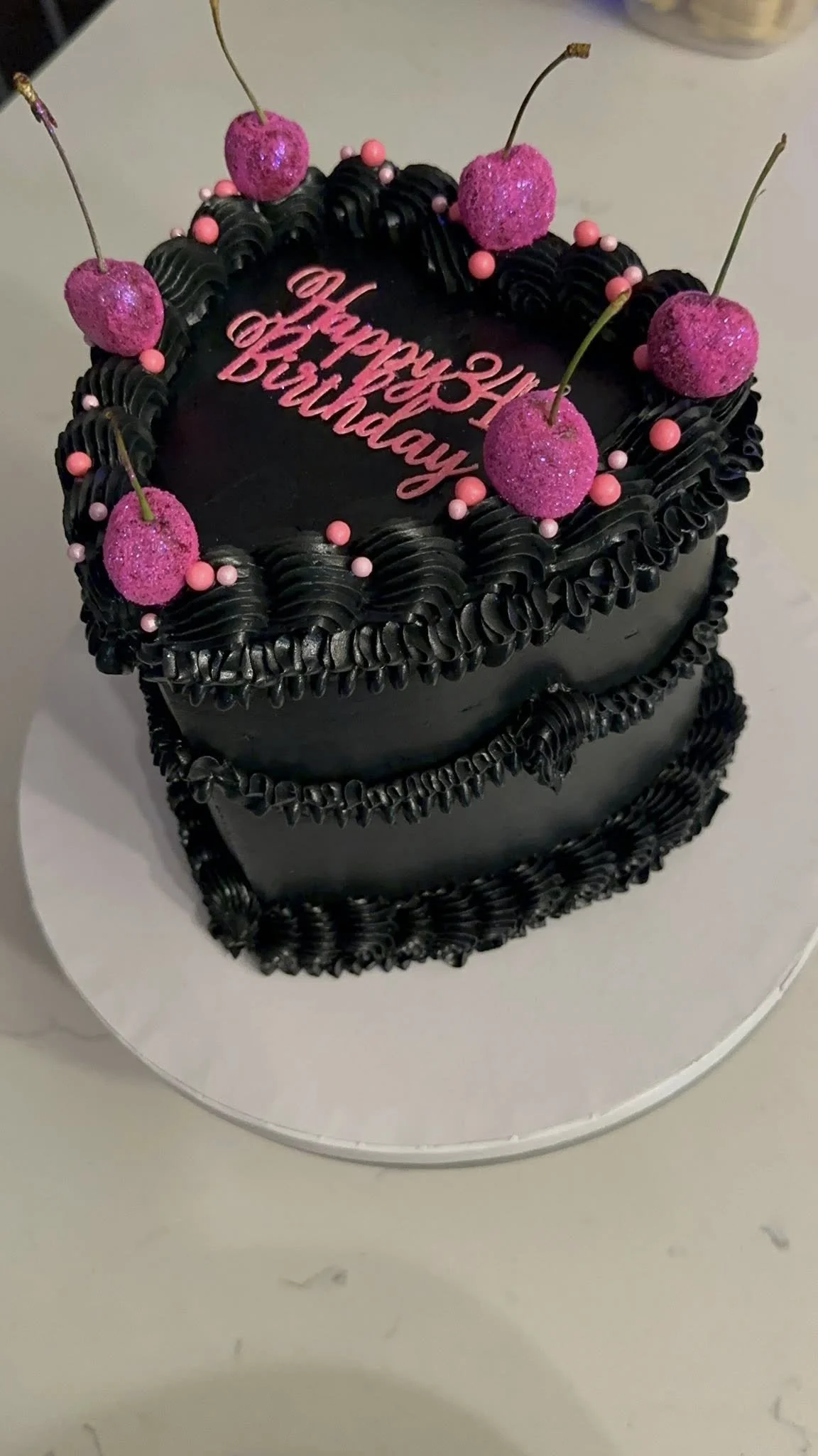A two-tier black birthday cake decorated with pink cherries and pink sprinkles, with a pink "Happy 24th Birthday" message on top.