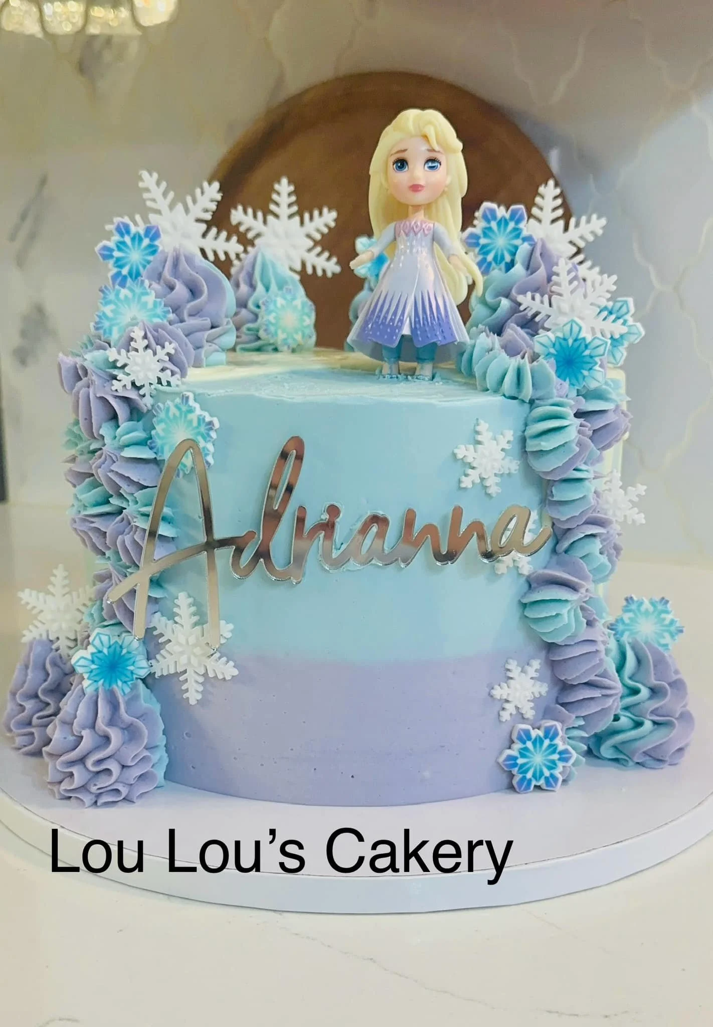 A pastel blue birthday cake decorated with snowflakes and a figurine of Elsa from Frozen on top. The cake has a gold 'Adriana' cake topper and is from Lou Lou's Cakery.
