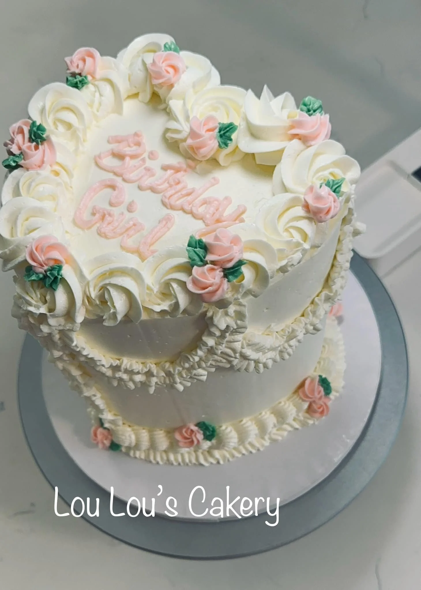 Two-tiered white birthday cake decorated with pink and green frosting flowers, with "Happy Birthday