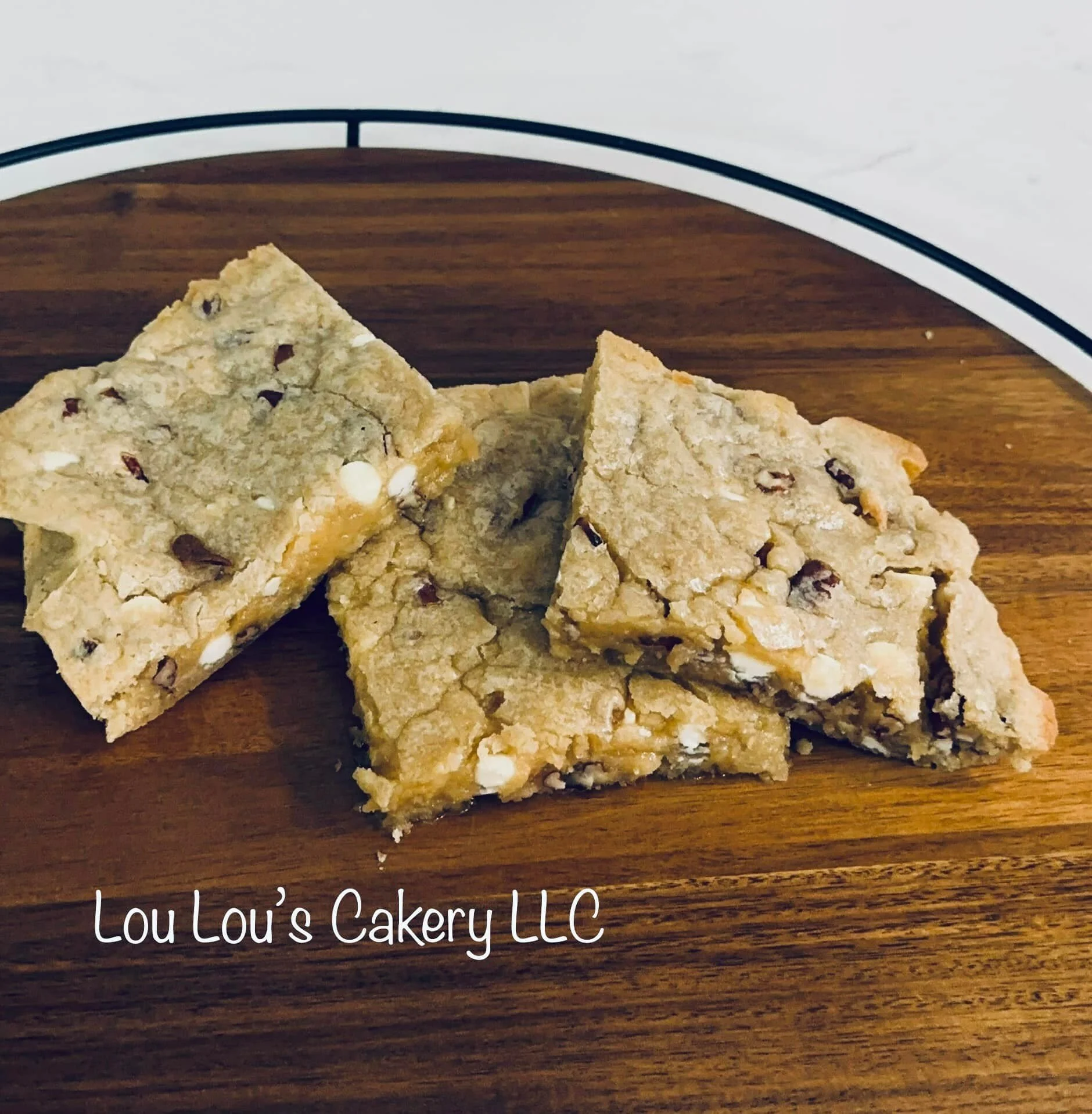 Four pieces of chocolate chip cookie bars on a wooden board with the text "Lou Lou's Cakery LLC" at the bottom.