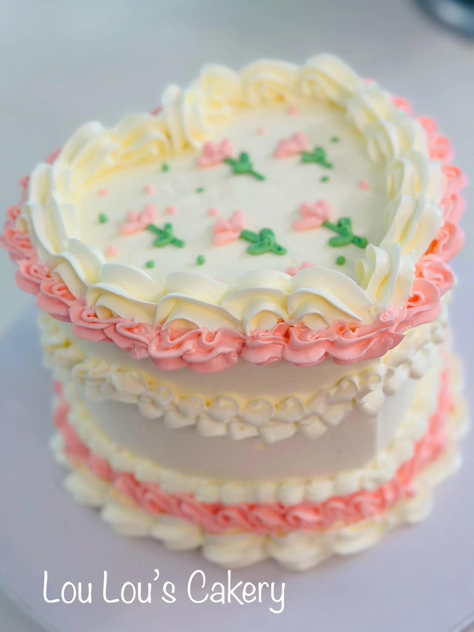 Decorative heart-shaped cake with white and pink frosting, topped with green and pink flower-shaped sprinkles, from Lou Lou's Cakery.