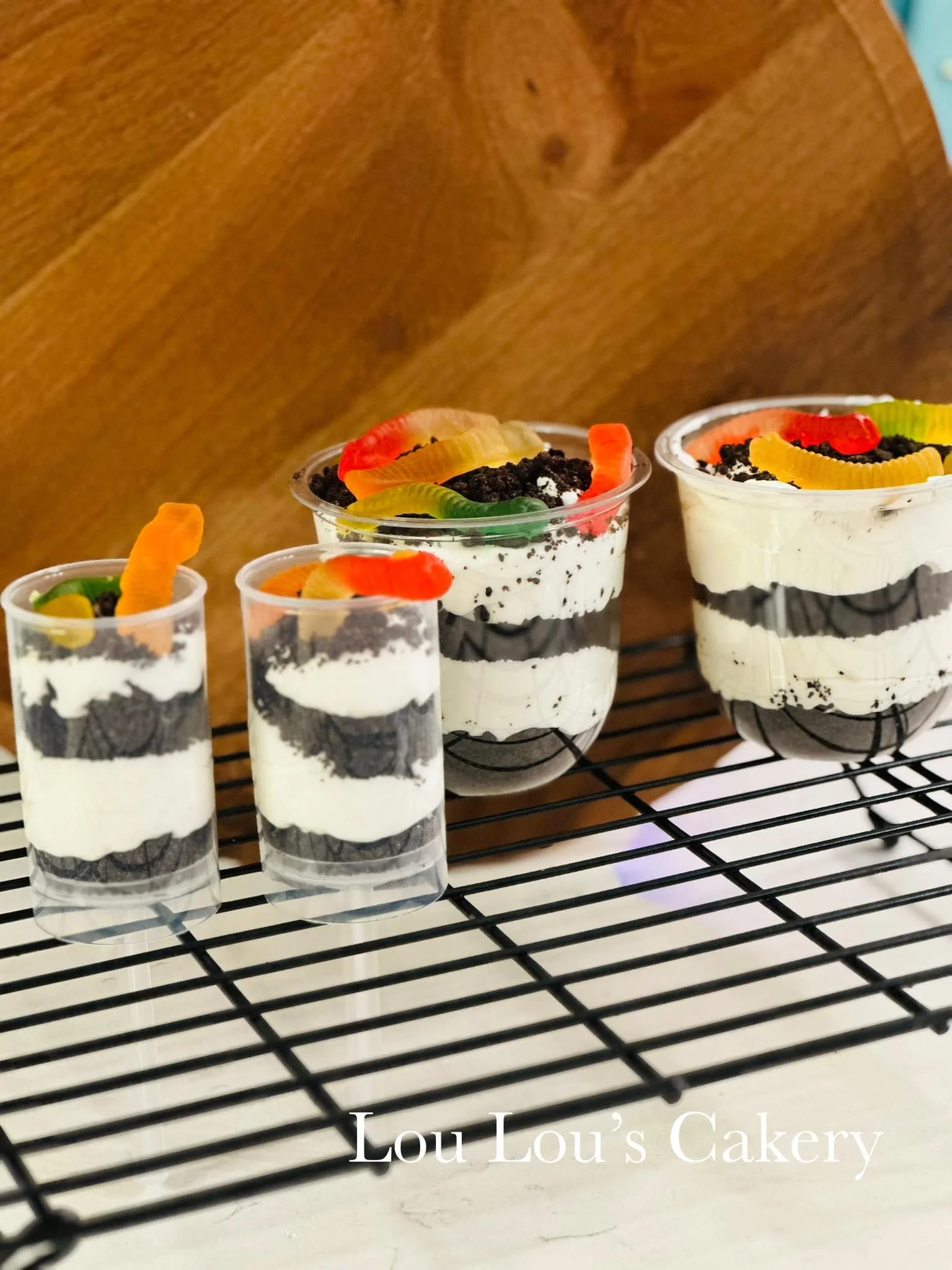 Four dessert cups with Oreo cookies, cream, and colorful gummy worm toppings placed on a cooling rack against a wooden background.