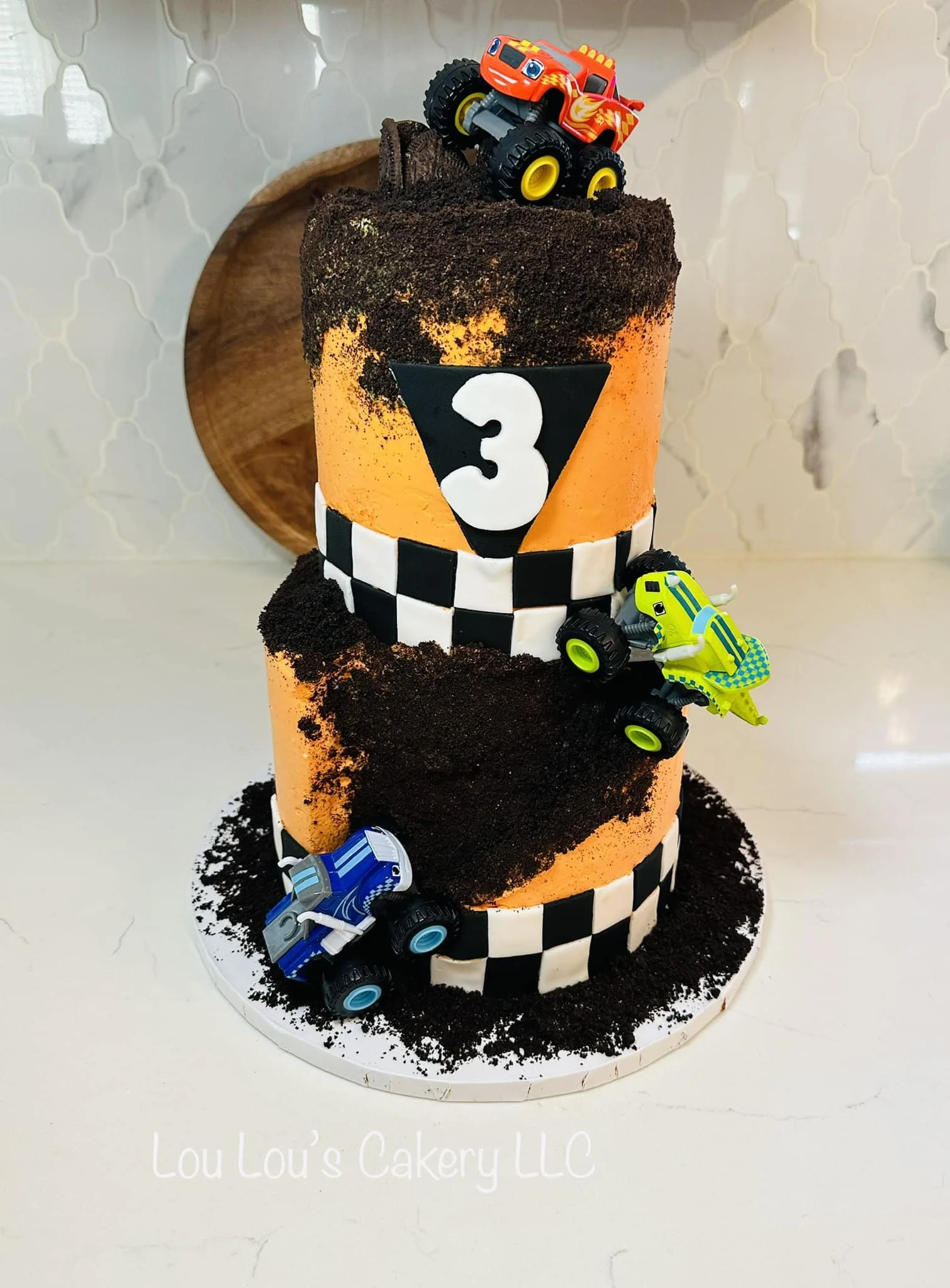 A two-tiered race car-themed birthday cake with orange and black coloring, black Oreos, and toy race cars on top and sides, decorated with checkered patterns and the number 3.