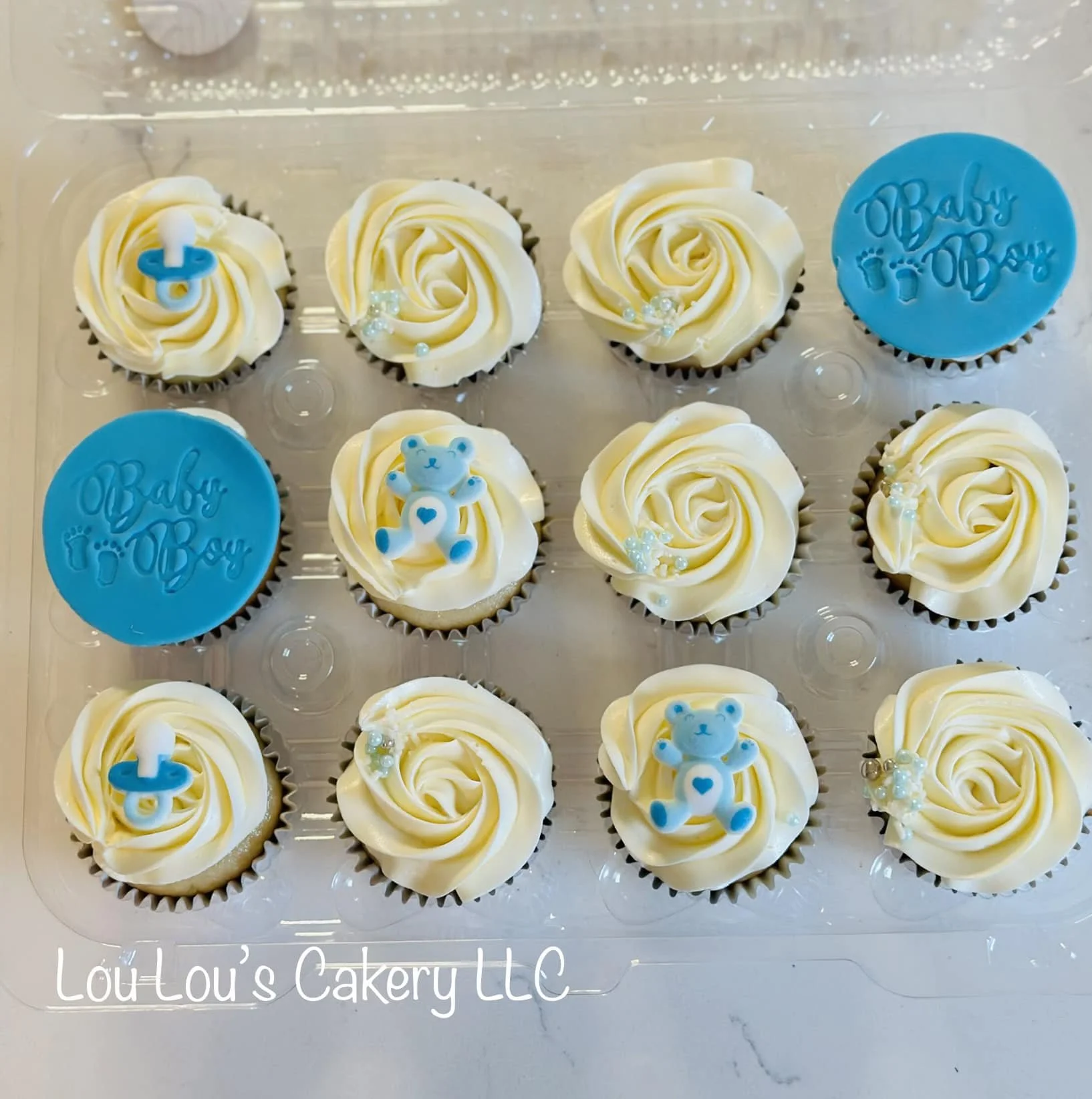 A tray of twelve baby-themed cupcakes, some topped with white frosting and small blue teddy bear or pacifier decorations, and others with blue fondant disks with "Baby Ot~e" written on them.