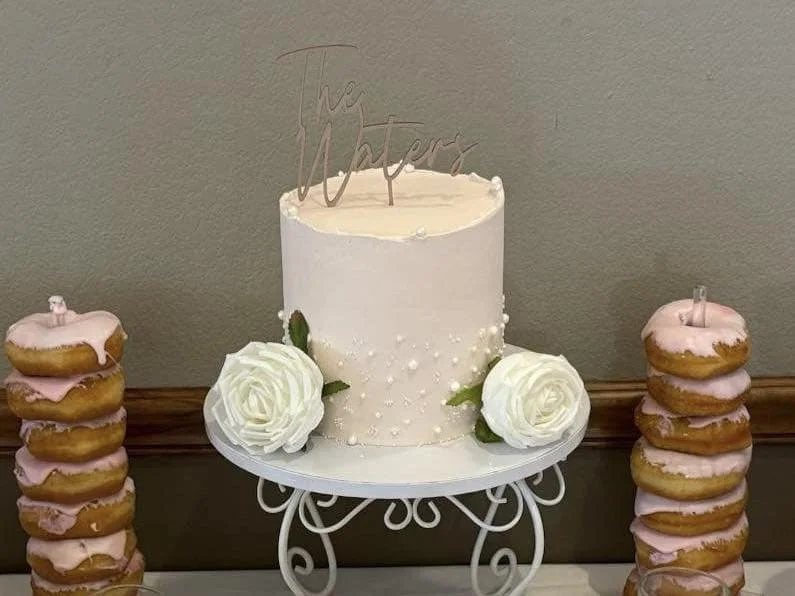 White birthday cake decorated with white roses and pearls, with a 'Mother' topper, flanked by stacks of donuts with pink icing.