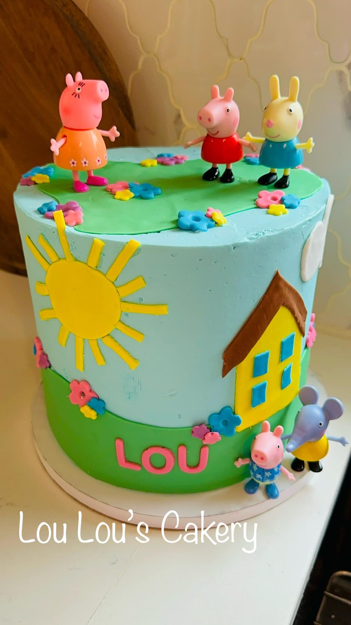 Colorful Peppa Pig themed birthday cake with figurines of Peppa Pig characters on top, decorated with a sun, house, and flowers, with the name 'Lou' on the side.